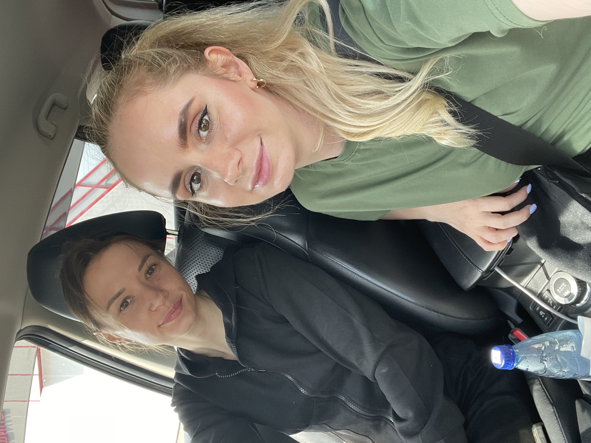 Two women are seen inside a car, captured in a selfie-style photograph during what appears to be daytime. The image is oriented vertically with the subjects looking towards the camera.
The woman in the foreground has blonde hair, styled with some strands pulled back, and is wearing a green short-sleeved top with a black seatbelt across her chest. She has prominent dark eyebrows, noticeable eyeliner, and subtle makeup, including light pink lipstick. She wears a small gold hoop earring in her visible ear and has light blue nail polish on her left hand, which is resting near her lap. She is smiling gently.
Behind her, in the passenger seat, is another woman with dark hair, which appears to be tied back. She is wearing a black zippered hoodie and also has a subtle smile, looking towards the camera.
The car interior features black leather seats and headrests. On the center console, between the two women, a part of the dashboard with controls is visible, including a knob with "4x4 AUTO" text. A silver cable is plugged in nearby. A plastic water bottle with a blue cap is resting on the console or floor area.
Outside the car window, through the bright daylight, some red and white structures are visible, possibly part of a building or an industrial area. Faint red Cyrillic text can be seen on a white background in the lower left window area, and also partially through the rear window, hinting at commercial or public signage. The overall mood conveyed by the women's expressions is relaxed and pleasant. The scene takes place in Kiparisovo-2, Russia.