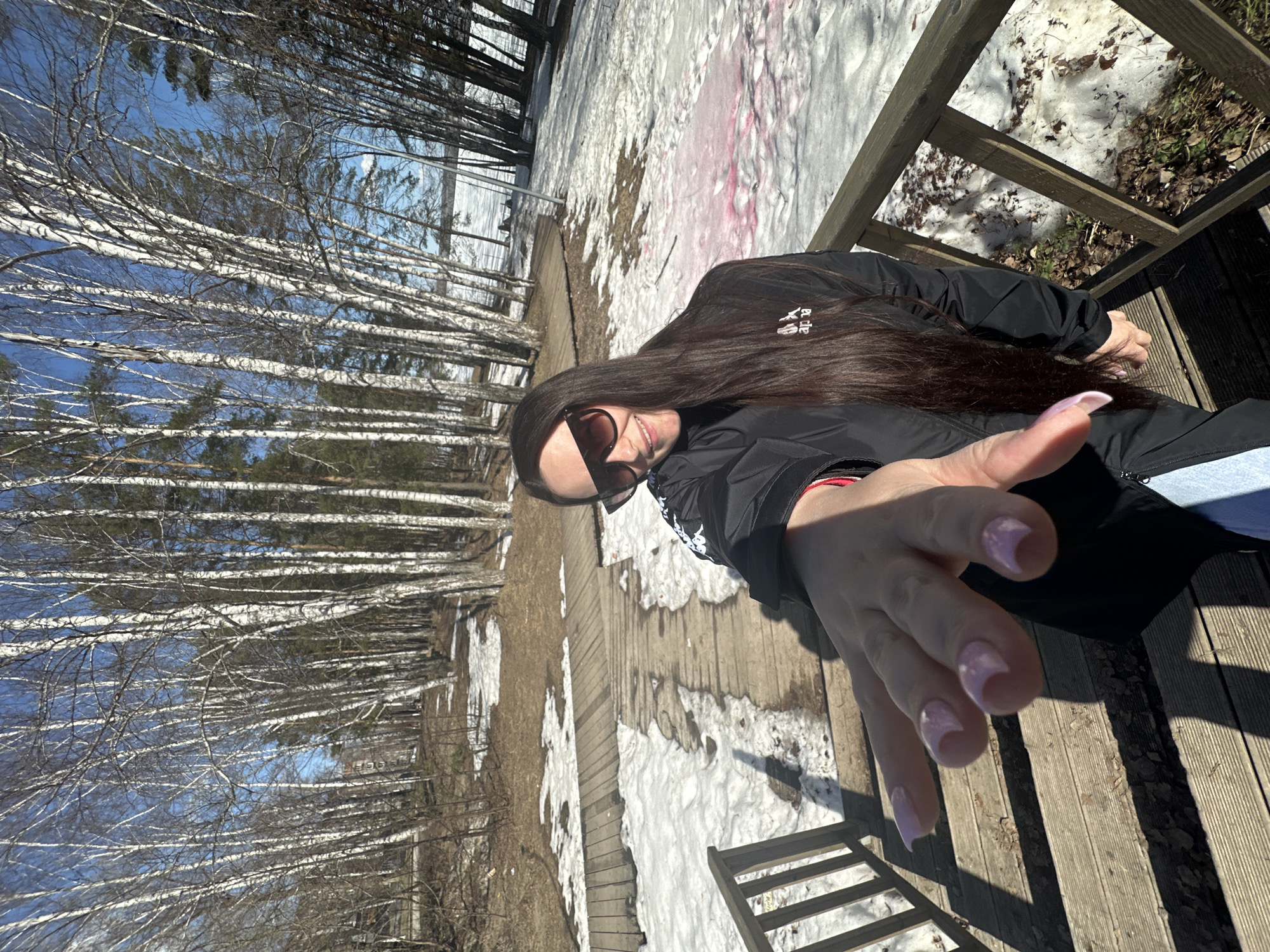 A woman is standing on a wooden deck or boardwalk in Chaikovsky, Russia. She is wearing sunglasses and a black jacket, and she is reaching out towards the camera with her hand. Her fingernails are light pink. The deck is partially covered in snow. There are many tall, thin birch trees on the left side of the image. In the background, there is a body of water.