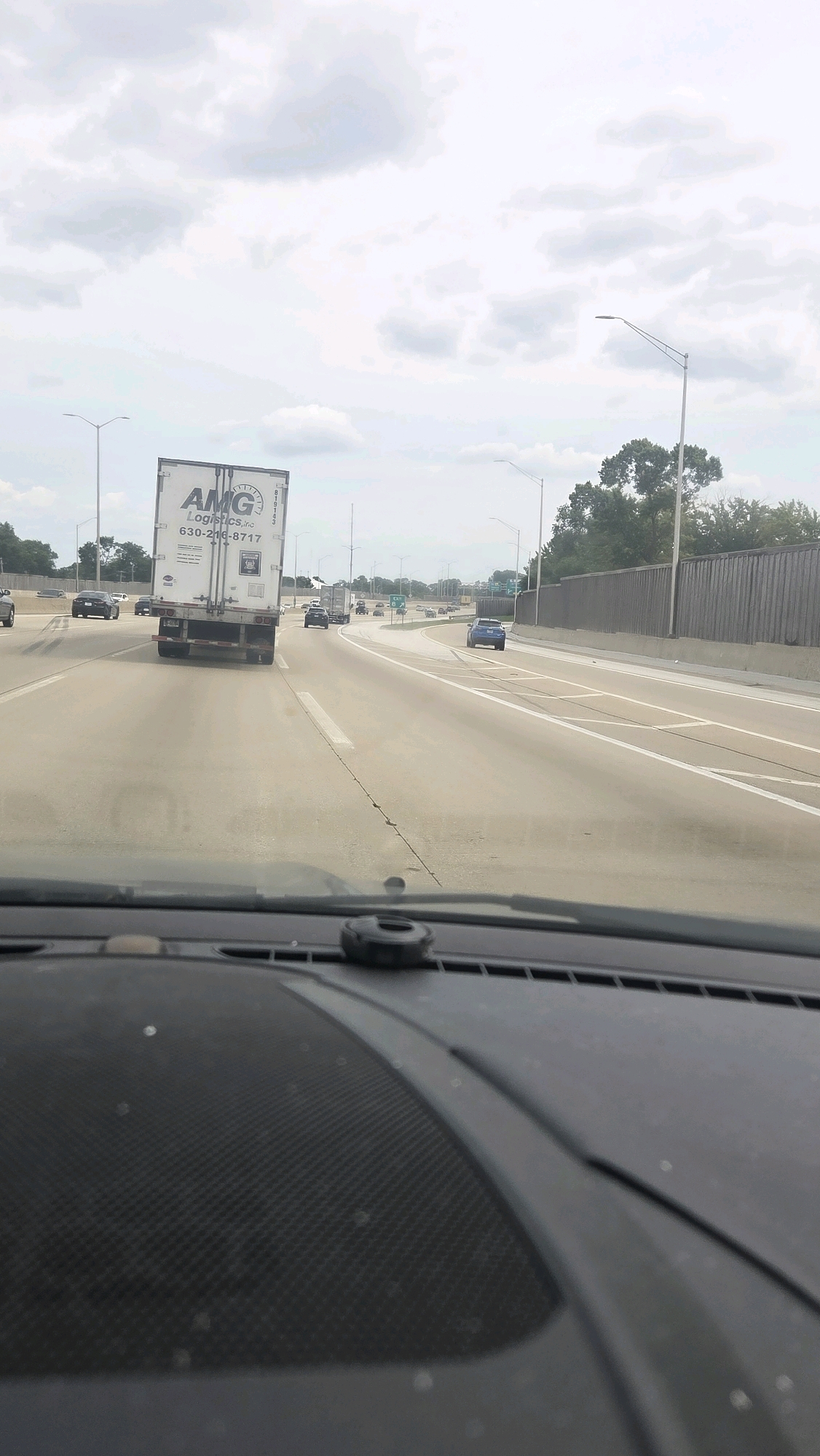 The image shows a view from inside a vehicle traveling on a highway. In the foreground, the dashboard of the car is visible, with a dark, textured surface and a small circular object mounted on it. The highway stretches ahead, with multiple lanes of traffic. Several vehicles are present on the road, including a white semi-trailer truck with "AMG Logistics, inc" and a phone number displayed on its side. Other cars and trucks are also visible in the distance. The highway is lined with trees and a fence on the right side. The sky is overcast with clouds. The scene appears to be within the city of Posen, Illinois, United States of America, based on the context provided by the crops.