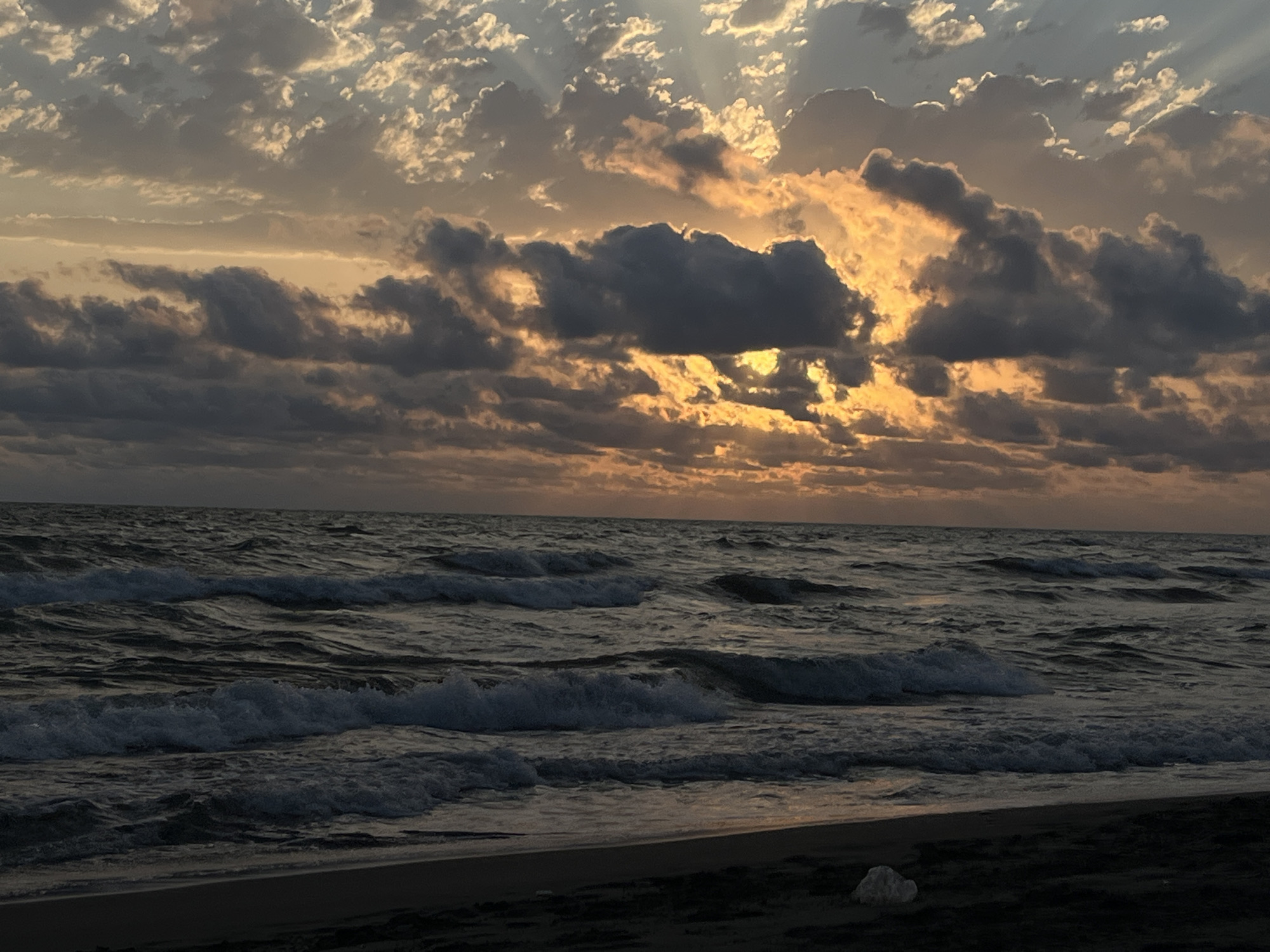 A wide-angle photograph captures a twilight scene at a beach in the village of Supsa, Georgia. The sky is dominated by dramatic clouds, with rays of sunlight breaking through, casting a warm orange glow in the upper half of the frame. Below the sky, dark, choppy waves crash onto a sandy shore. The water is a deep, dark blue-gray, reflecting the subdued light. The foreground shows a dark, sandy beach with a few scattered white objects, possibly shells or stones, one prominent white object rests on the sand near the waterline. The horizon line is clearly defined, separating the turbulent sea from the vibrant, cloudy sky. The overall impression is one of a serene yet powerful natural landscape at dusk.