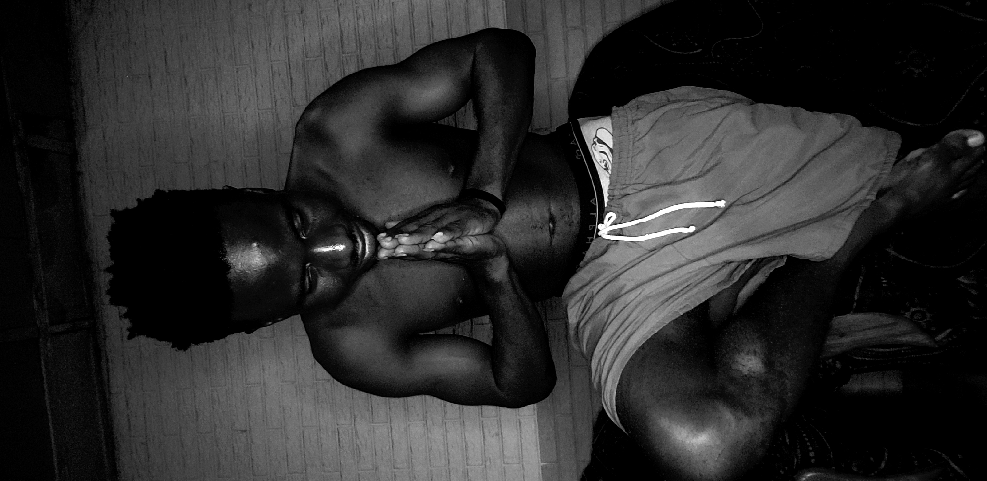 A shirtless, dark-skinned man is lying on his back, facing the camera. He is wearing shorts with white drawstrings. His hands are clasped together near his chin. The environment appears to be a room with a tiled wall in the background. Based on the location information, the room is in Narayi, Nigeria. The scene is lit dimly.