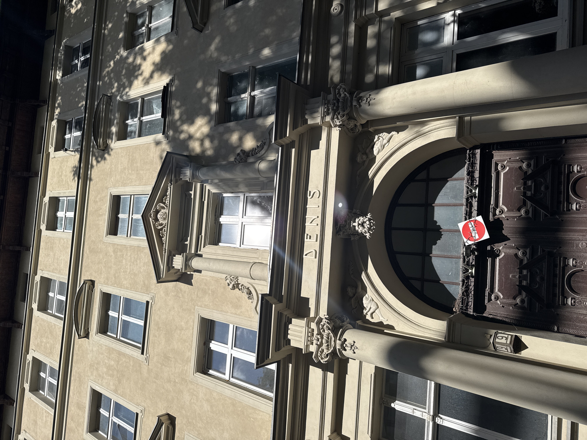 This is a low-angle, close-up, outdoor shot of the facade of an old building in Poznan, Poland. The building appears to be several stories high with numerous windows. The facade is a light tan color and is adorned with decorative architectural elements such as columns, pediments, and carvings. Above the arched doorway, the word "DENIS" is carved into the building. On the dark wooden double doors of the entrance, a small, circular red and white sign with a black line through it is affixed. Sunlight casts strong shadows across the building, suggesting it is daytime.