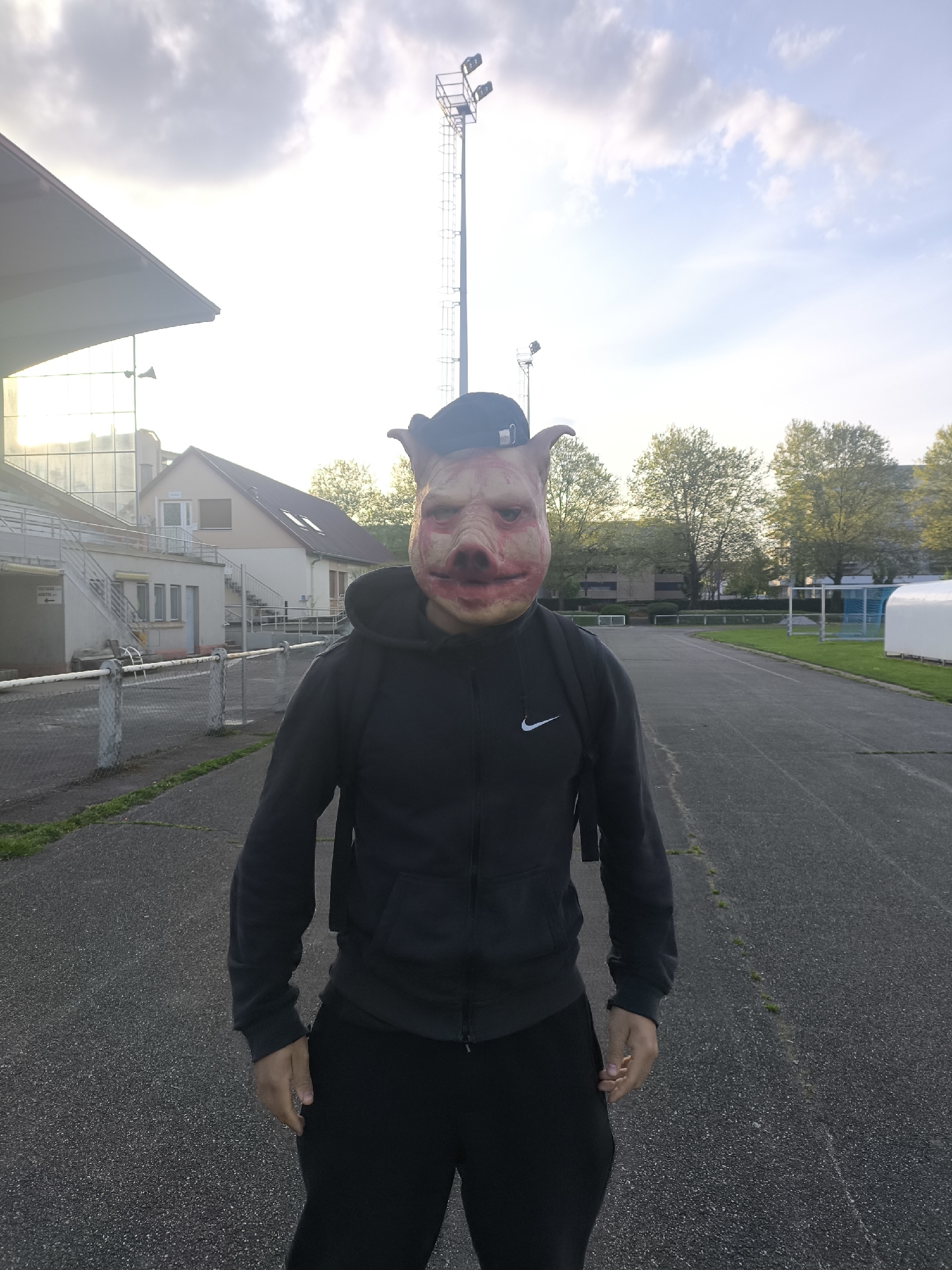 The photo shows a person wearing a pig mask and a black hoodie with a white Nike swoosh, standing on an athletic track. The person is also wearing black pants and a black baseball cap. A backpack is slung over their shoulders. The setting appears to be an athletic field or stadium in Schiltigheim, France. In the background, there is a stadium seating area, buildings, trees, and a tall light pole. The sky is cloudy with some sunlight breaking through. The track is paved with asphalt.