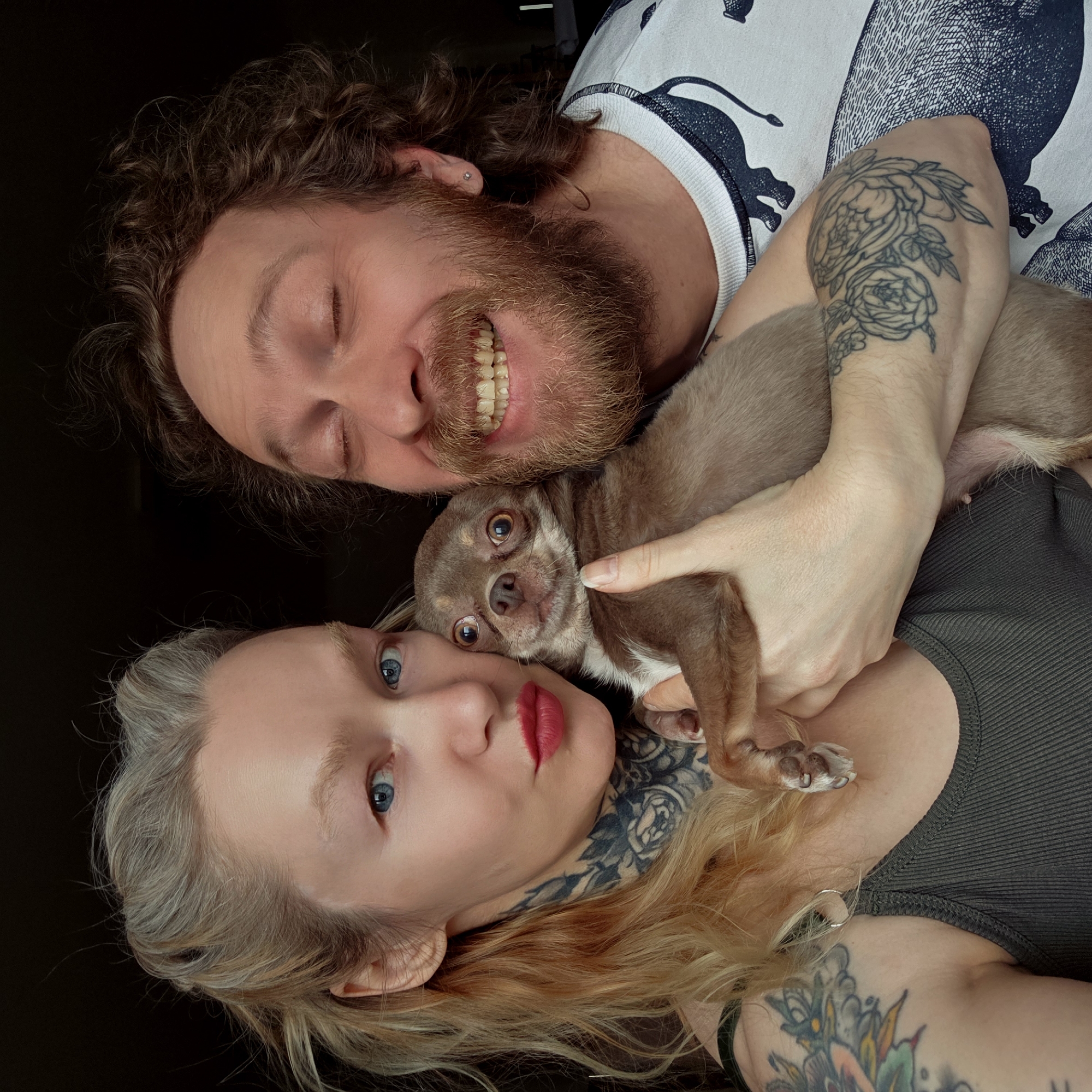 A high-angle, rotated portrait captures a man, a woman, and a small dog posing closely together against a dark background, likely while lying down. The man is positioned above the dog, and the woman is below the dog. Their heads are oriented towards the left of the frame.
The man, with curly brown hair and a full beard, smiles widely, revealing his teeth. He has a small piercing in his left ear. He wears a white sweatshirt adorned with a dark, patterned design, possibly depicting animals. A floral-patterned tattoo is visible on his right arm, which is wrapped around the dog.
Below the man, a small, light brown Chihuahua with large, dark eyes looks directly at the camera, nestled between the two people. The man's hand gently holds the dog, and its small paws are visible.
The woman has blonde hair with darker roots, striking blue eyes, and bright red lipstick. She maintains a more composed expression while looking towards the camera. Prominent tattoos are visible on her: a detailed floral design on her neck/shoulder area, and a colorful, intricate pattern on her left arm/shoulder. She is wearing a dark, ribbed tank top or camisole.
The image conveys a sense of warmth and affection shared among the subjects. There is no visible text or discernible time of day or outdoor location cues.