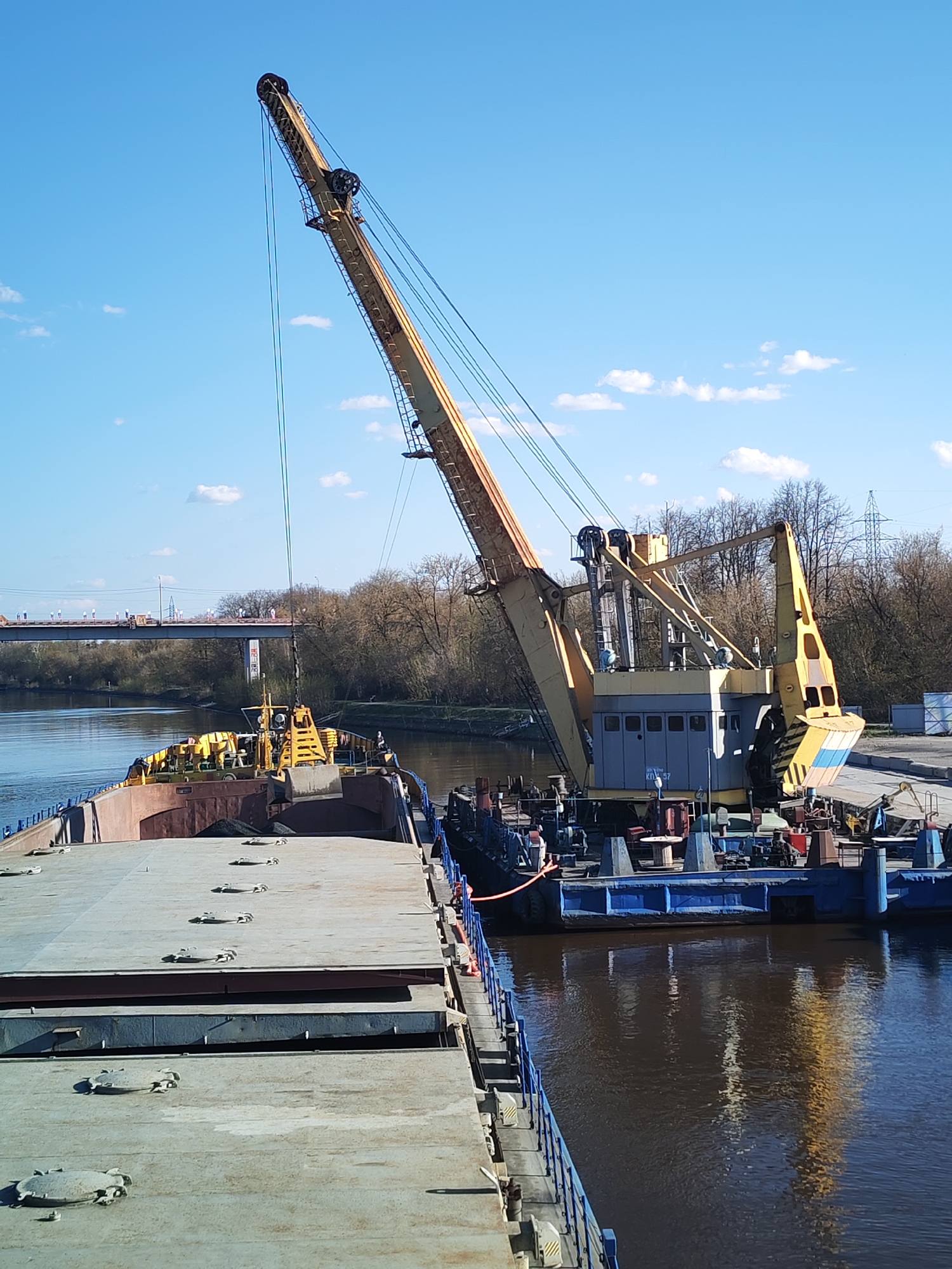 The image depicts a river scene in Yakhroma, Russia, dominated by an industrial operation involving barges and a crane. A large yellow crane mounted on a blue barge is prominent on the right side of the frame. The crane's boom is extended upwards at an angle. A second barge, partially filled with a dark material, is docked next to it. On the left, another barge stretches into the foreground. The river reflects the crane and sky. In the background, there is a bridge, trees, and power lines. The sky is blue with scattered white clouds. No people are clearly visible in the image.