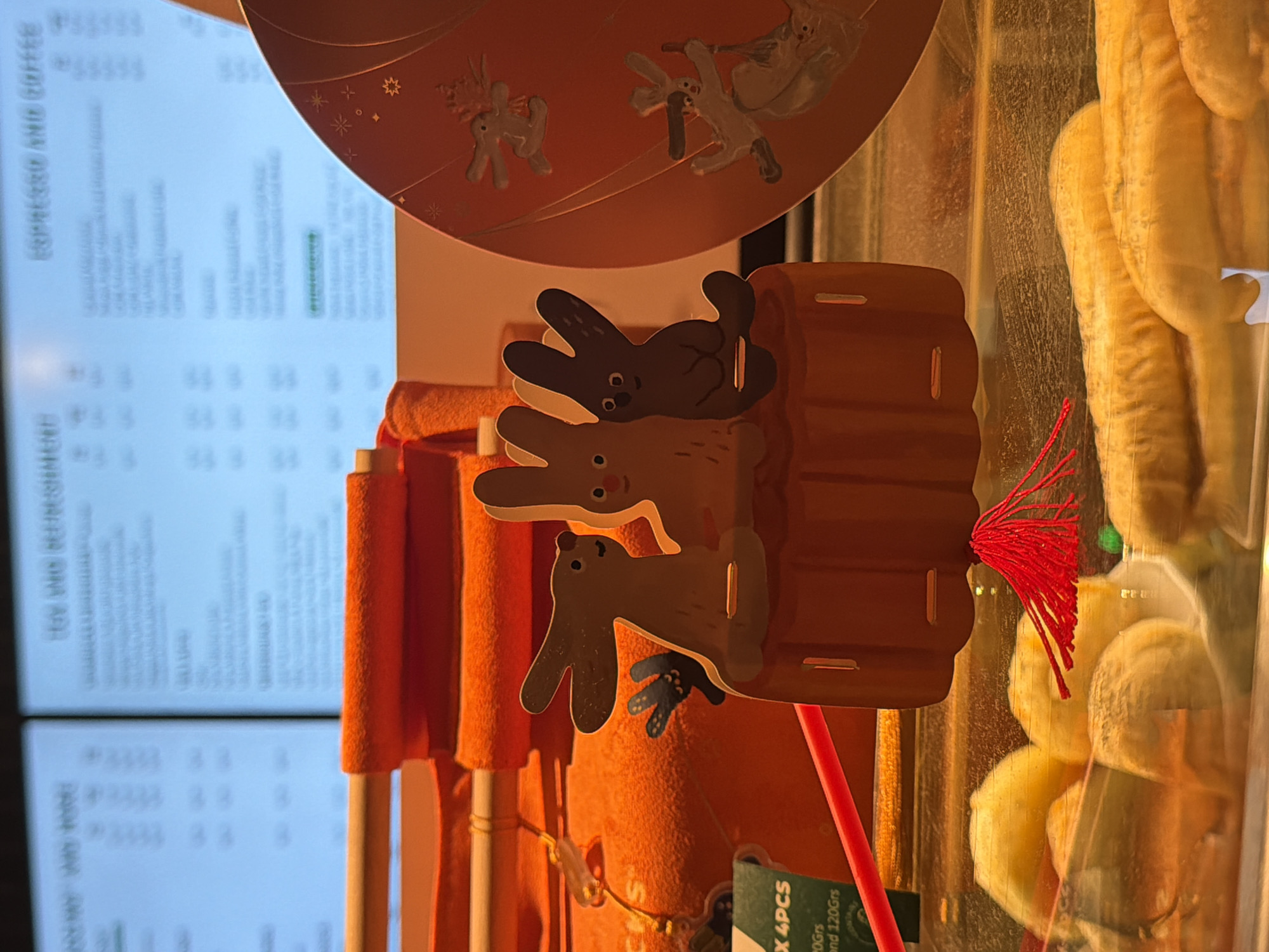 The image is a close-up shot of a display case in what appears to be a cafe or bakery. The display case contains several yellow, bread-like pastries. In the foreground, a decorative cardboard cutout of three cartoonish rabbits sits on an orange, pumpkin-shaped base. A red tassel hangs from the base. To the right, a circular orange decoration features more cartoon rabbits. Behind the display, two large digital screens show menu items. The screen on the left has a grid of text and prices, while the screen on the right displays "ESPPRESSO AND COFFEE" and a list of items. A portion of an orange towel or cloth is visible behind the rabbit cutout. The overall impression is a festive or seasonal display, possibly for an event like Easter, given the rabbit theme. The scene is brightly lit, with warm tones dominating the color palette due to the orange decorations and the lighting. There are no people visible in the image. The location is stated to be in the city of Da Nang, Vietnam.