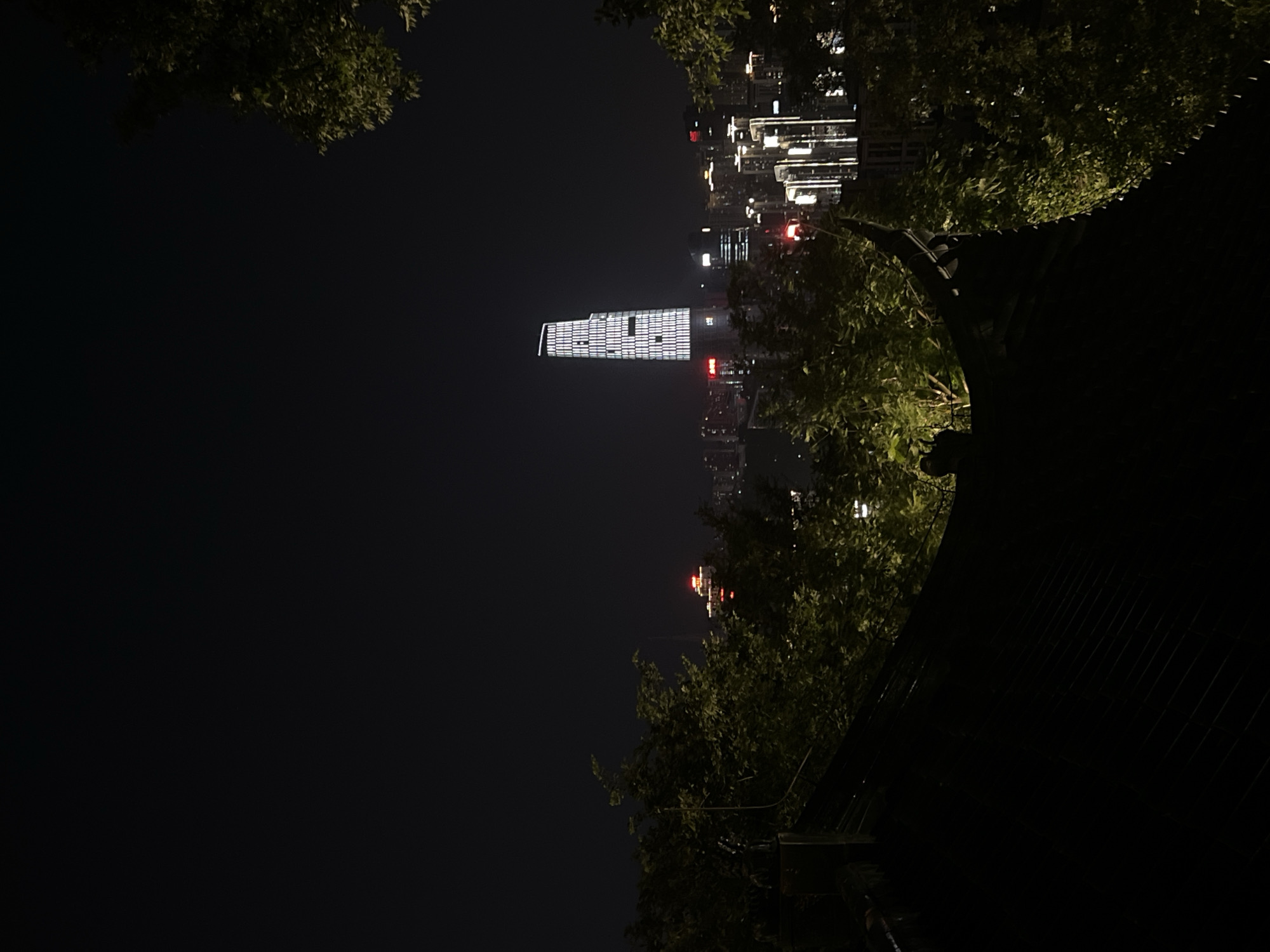 This nighttime photograph captures a cityscape, specifically the Liangjiang New Area in Chongqing, China. In the foreground, the dark silhouette of a traditional Chinese architectural element, likely a roofline with decorative eaves, dominates the lower portion of the frame. Lush green foliage of trees frames the scene from the left and right, partially obscuring the view of the city beyond.
The middle ground is a tapestry of urban lights. Numerous buildings, varying in height and illuminated by a multitude of lights, create a vibrant urban sprawl. Many of these structures are modern high-rises, their windows glowing warmly against the dark sky.
A prominent, brightly lit skyscraper stands out in the center of the image. This building, characterized by its distinctive white lattice pattern of lights, is a focal point of the composition. Other buildings in the distance also display illuminated windows and architectural lighting. A building with prominent red lighting is visible behind the trees.
The overall environment is that of a developed urban center at night, with a clear contrast between the dark, natural elements in the foreground and the illuminated, man-made structures in the background. There are no discernible people or specific actions depicted in the image.