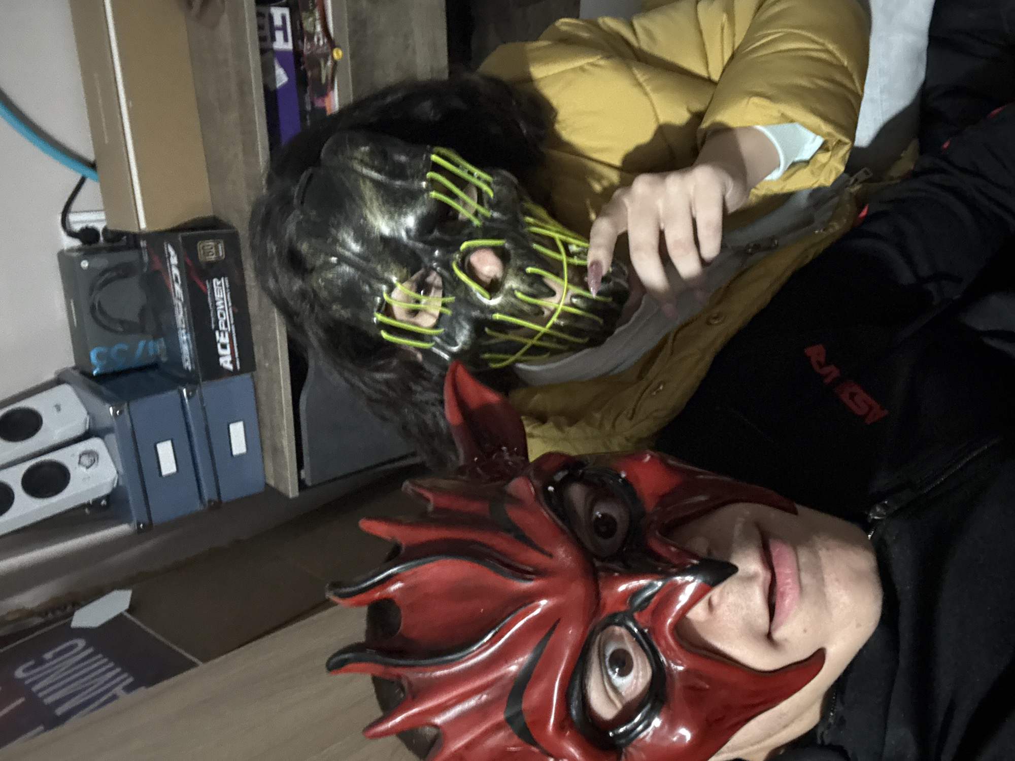 Two people wearing masks are visible. One person wears a red devil-like mask. The second person wears a black mask with yellow neon-like detailing. The person in the red mask is wearing a black jacket. The person in the black mask is wearing a yellow puffer jacket. The location is not specified in the image. The location of Goritsa, Bulgaria, cannot be determined from the information provided.