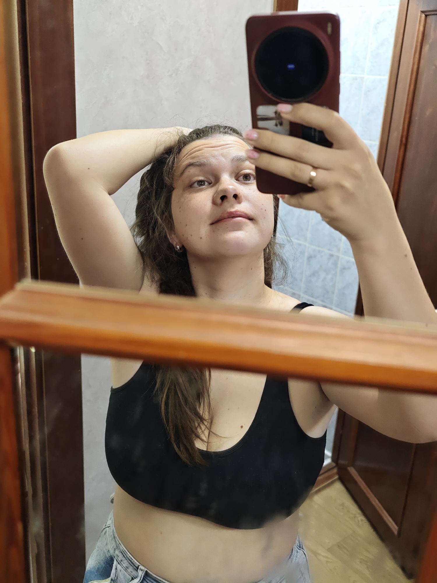 A woman is taking a selfie in a mirror. The woman has fair skin with freckles, brown hair, and is wearing a black tank top and blue jeans. She is holding a red smartphone with a large circular camera lens. The background shows a bathroom with white and blue tiled walls and dark wooden doors. The reflection in the mirror is of the woman and the bathroom. The location is not specified and there is no indication this image was taken in Chaikovsky, Russia.