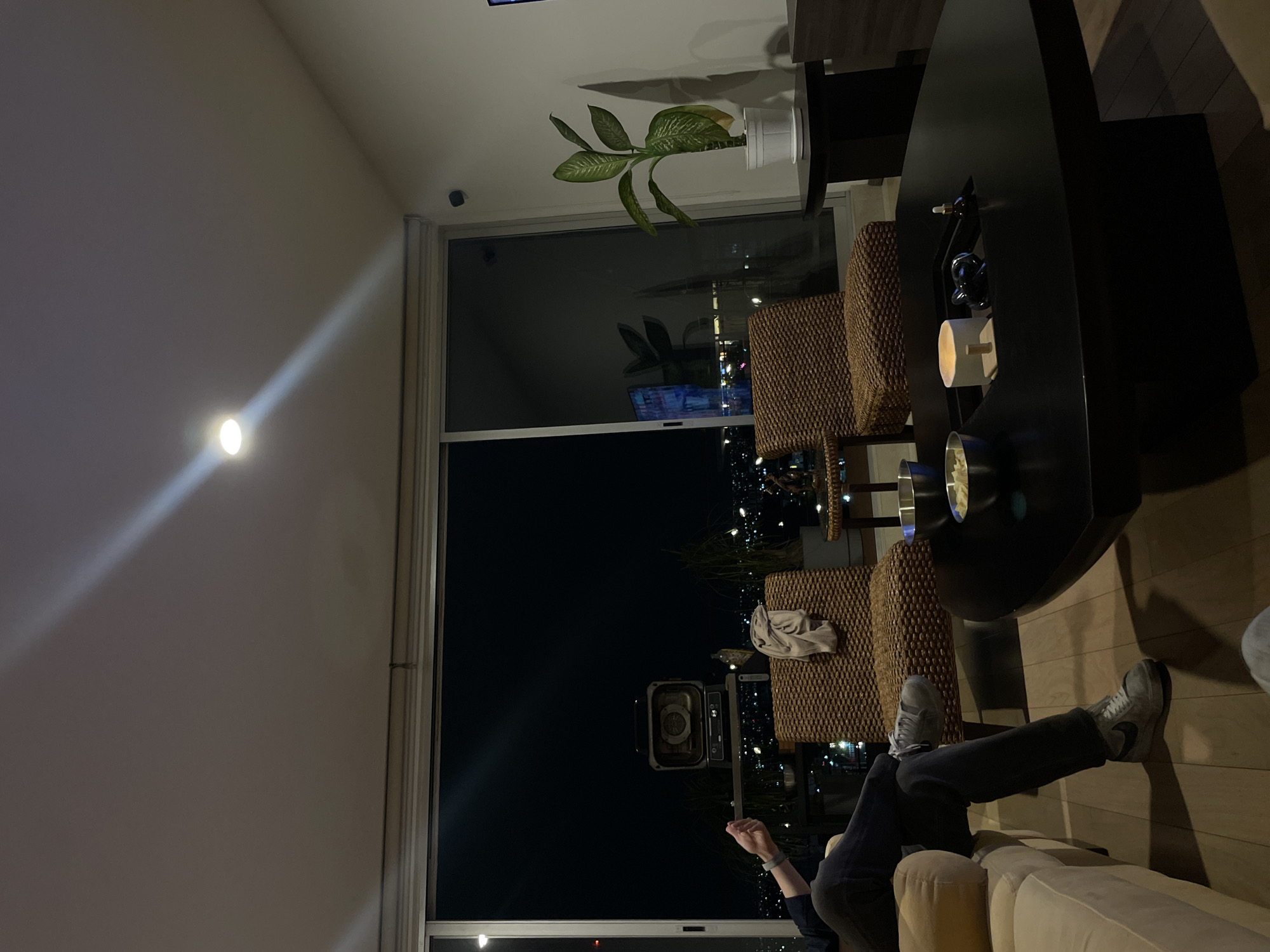 Here is a forensic description of the image:
**Setting:** An interior living room with a large window overlooking a city skyline at night. The room has a vaulted ceiling with a recessed light fixture.
**People:** One individual is visible, reclining on a beige sofa. The individual is wearing dark pants and grey athletic shoes. The person's hand and arm are partially visible.
**Objects:** The room is furnished with two wicker chairs and a black, modern coffee table. The coffee table holds decorative items, including a candle and dark glass objects. A potted plant sits near the window. The window offers a view of the city lights.
**Actions:** The individual is reclining on the sofa with their legs extended.
**Environment:** The cityscape visible through the window suggests an urban setting, though the exact location in Puebla, Mexico, cannot be definitively determined from the image. The overall lighting is dim, with the primary light sources being interior fixtures and the city lights seen through the window.