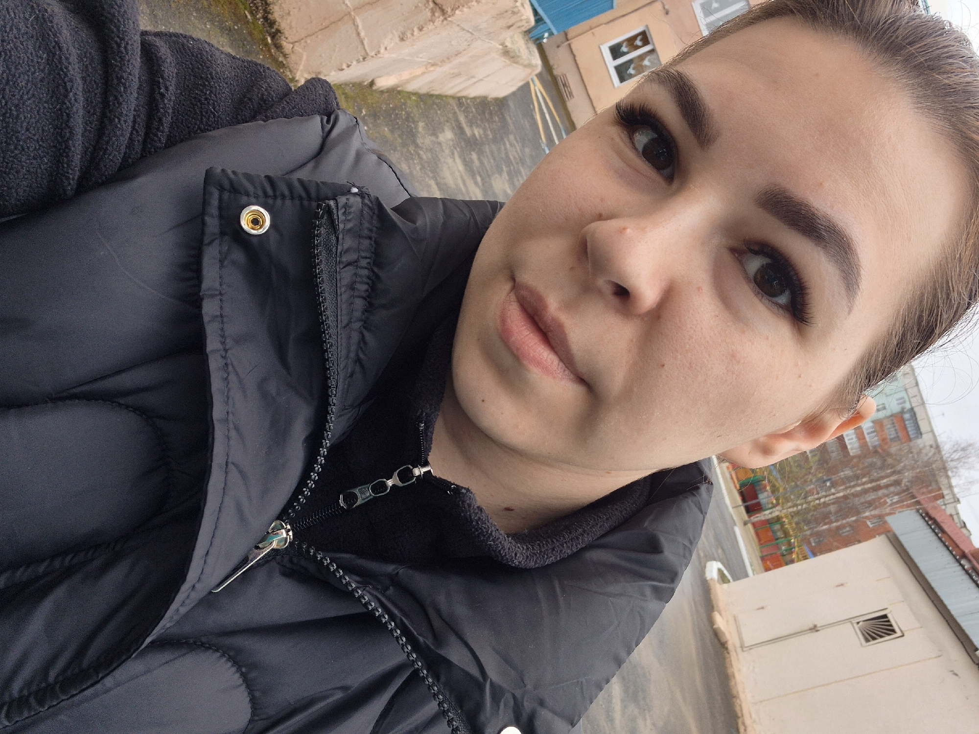 A woman is taking a selfie. She is wearing a dark-colored puffer jacket. The background shows a cityscape, identifying the location as Tobolsk, Russia. Buildings and possibly trees are visible in the background.