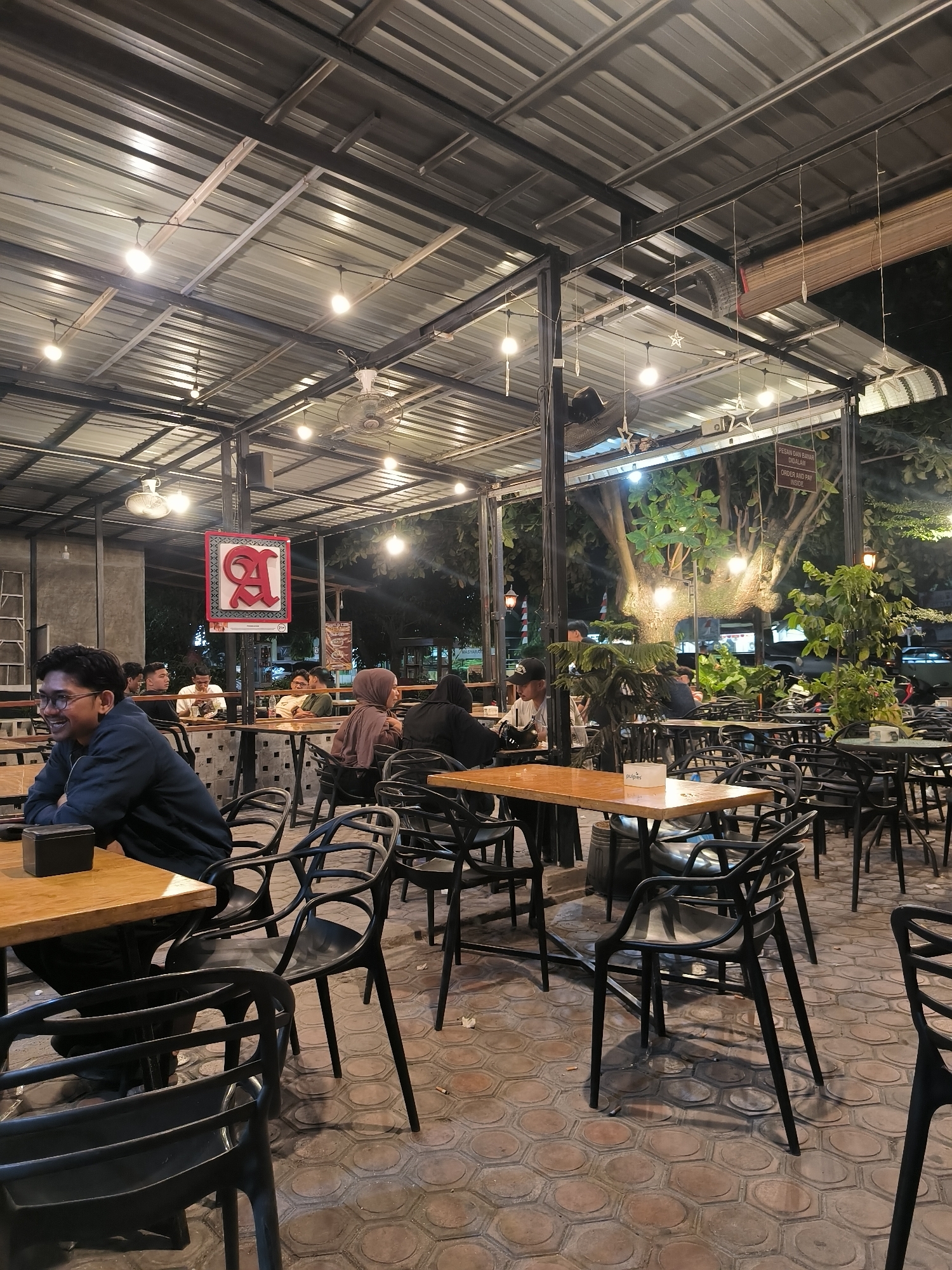 The image depicts an outdoor seating area of a restaurant or cafe in Lhokseumawe, Indonesia. The area is covered by a corrugated metal roof supported by metal beams, with hanging lights illuminating the space. Several tables with chairs are arranged on a patterned brick floor. People are seated at various tables, engaged in conversations or looking at their phones. In the foreground, a man in a dark jacket and glasses is seated at a wooden table, looking to the left. Behind him, other patrons are visible, some with food and drinks. A sign with a stylized red letter 'A' is hanging prominently in the middle ground. Lush green trees and foliage are visible in the background, suggesting an urban park or garden setting. The overall atmosphere appears relaxed and social.