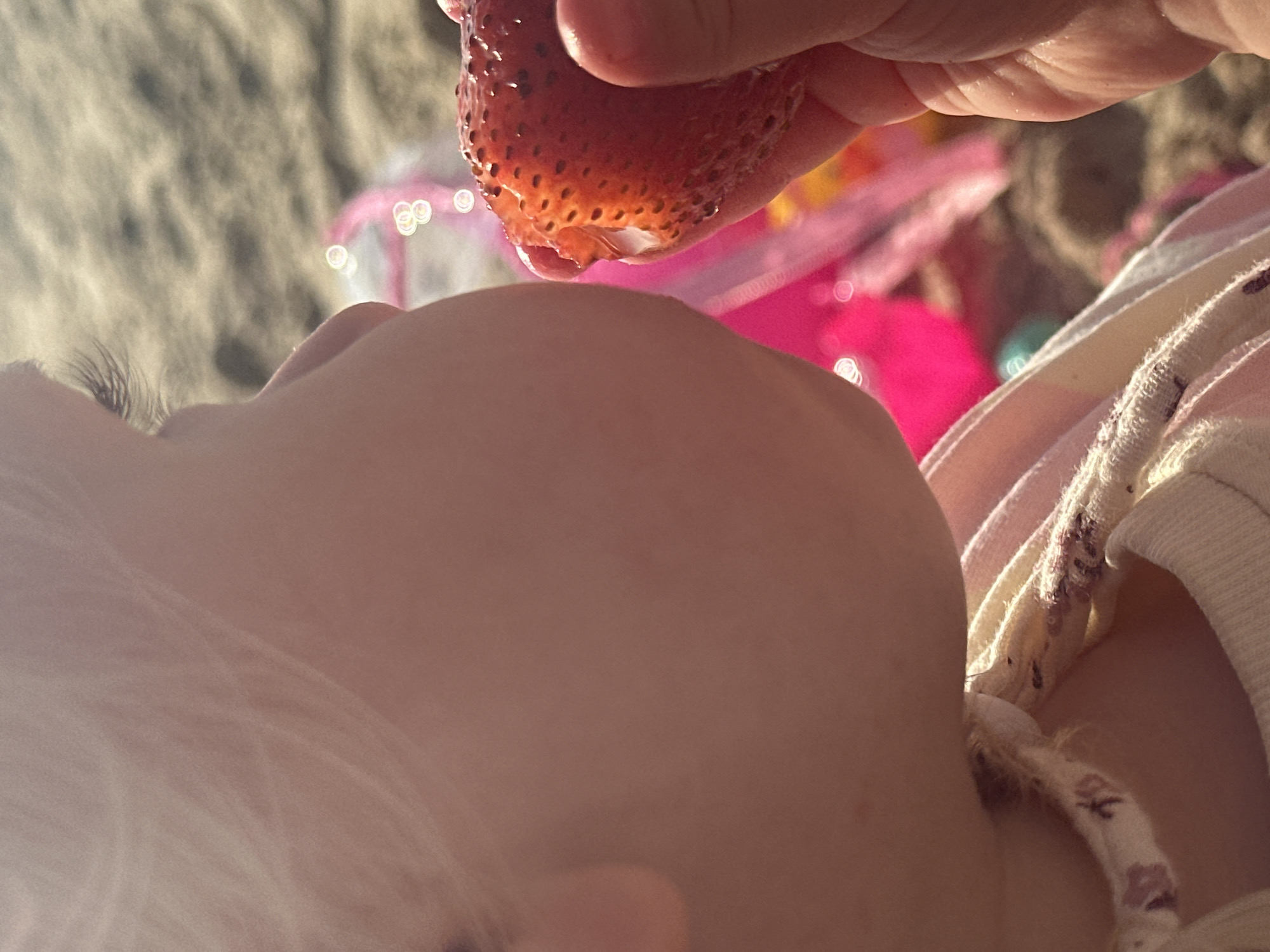 A low-angle, close-up shot captures a child's face as someone holds a strawberry above them. The child's head is tilted back, with light blonde hair and eyelashes visible. The strawberry is bright red with seeds and appears to be dripping juice. Behind the child and strawberry, there is a blurred background of what appears to be sand, possibly from a beach, and colorful plastic toys. The person holding the strawberry is only partially visible, showing their fingers. There is a fabric with a floral pattern visible as well, likely part of the child's clothing. Without more information, identifying the exact location in Tel Aviv, Israel is impossible.