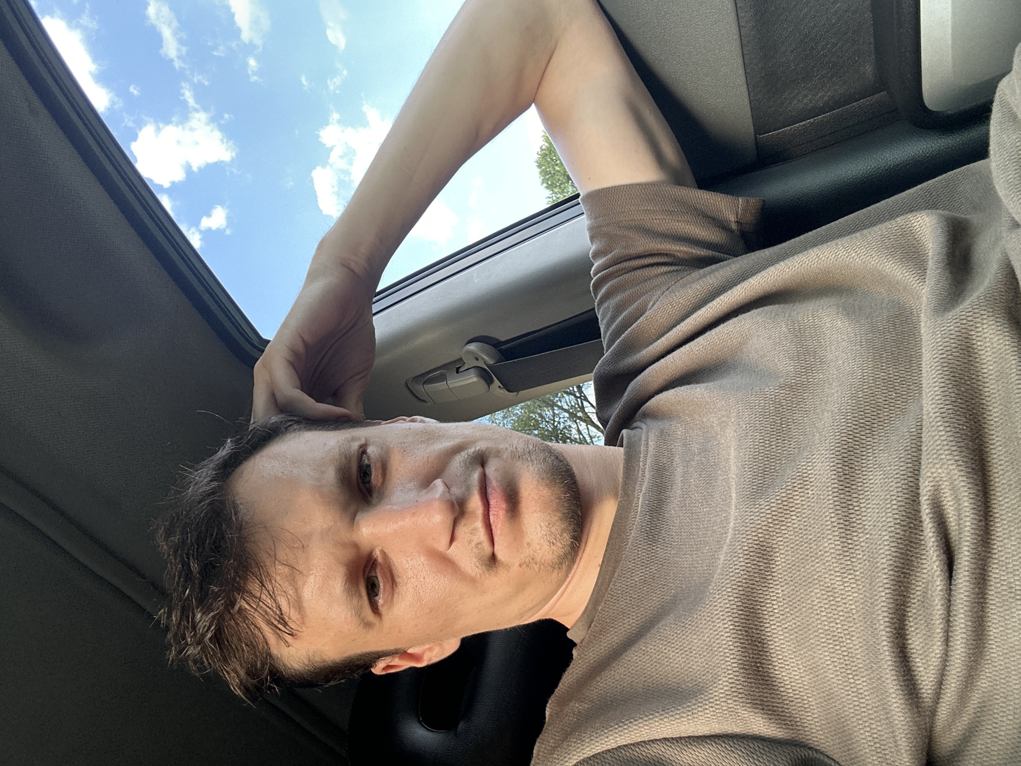 The image is a close-up selfie of a man with short, dark hair and light skin, taken inside a vehicle. He is wearing a brown t-shirt with a waffle knit texture. His right arm is bent, with his hand resting on the back of his head, near his temple. He is looking directly at the camera with a neutral expression. The car interior is visible, including a dark seat, a seatbelt, and a portion of the window frame. Through the open window, a bright blue sky with scattered white clouds and green trees can be seen. The lighting suggests it is daytime, possibly late afternoon due to the warm tones. The man appears to be in a relaxed pose. No specific location in Makiv, Ukraine is identifiable from the image.