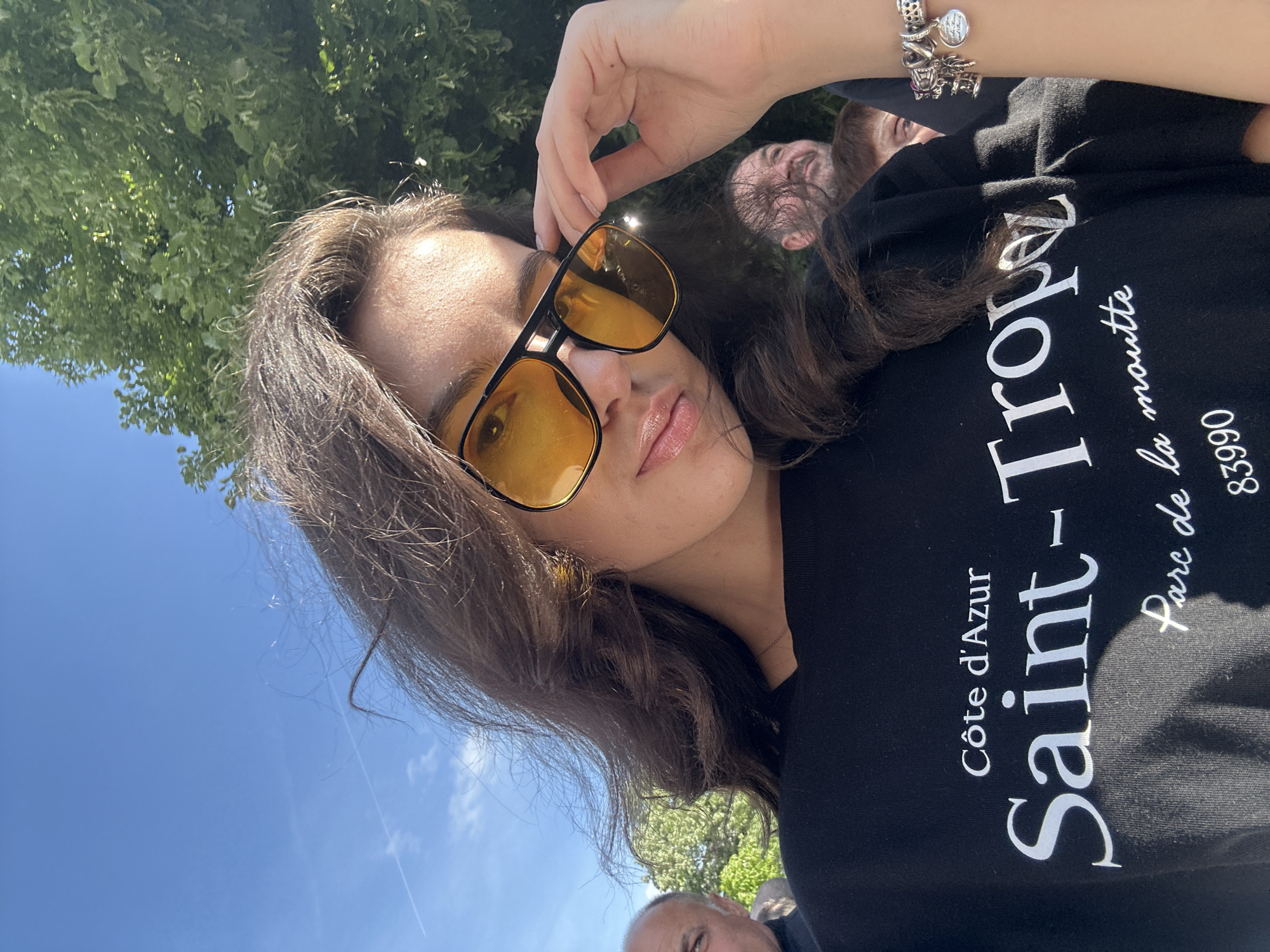 A woman in Saint-Tropez is taking a selfie in front of a tree with another woman and a man visible in the background. She is wearing sunglasses with amber lenses, a black t-shirt with white text reading "Cote d'Azur, Saint-Tropez, Parc de la moutte, 83990", and a bracelet with multiple charms on her left wrist. The sky is visible in the background, with a contrail running across.