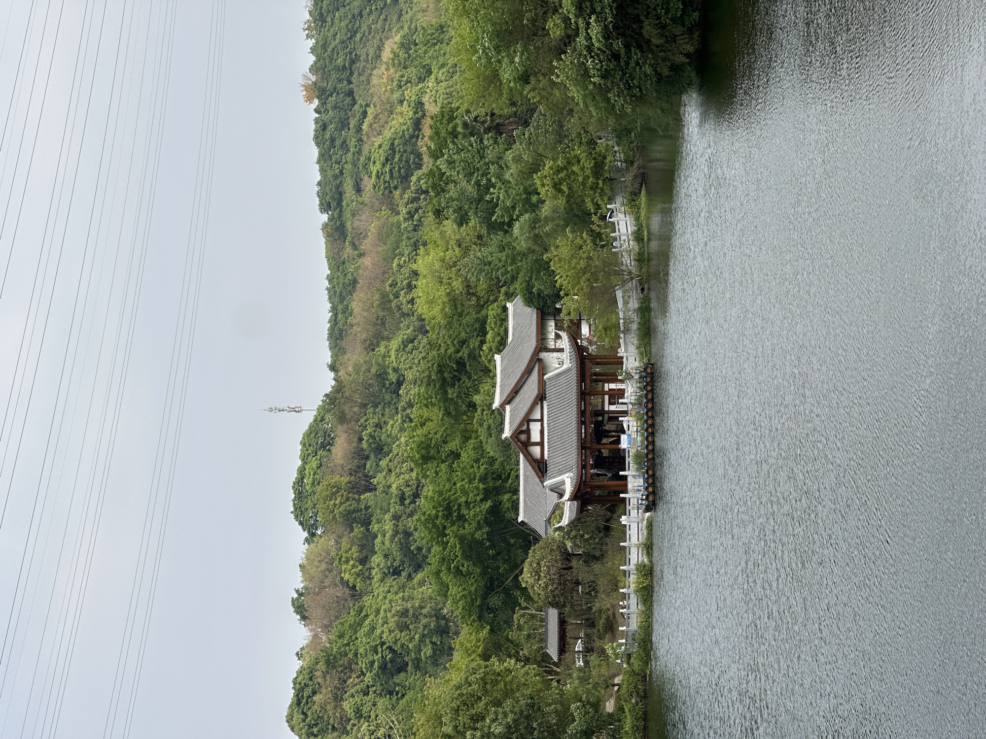 A landscape photo shows a body of water to the right with a green hillside to the left. A traditional-style building with dark tiled roofs is nestled between the water and the trees. Power lines are visible in the sky. The city of Liangjiang, China is not discernible from the image.