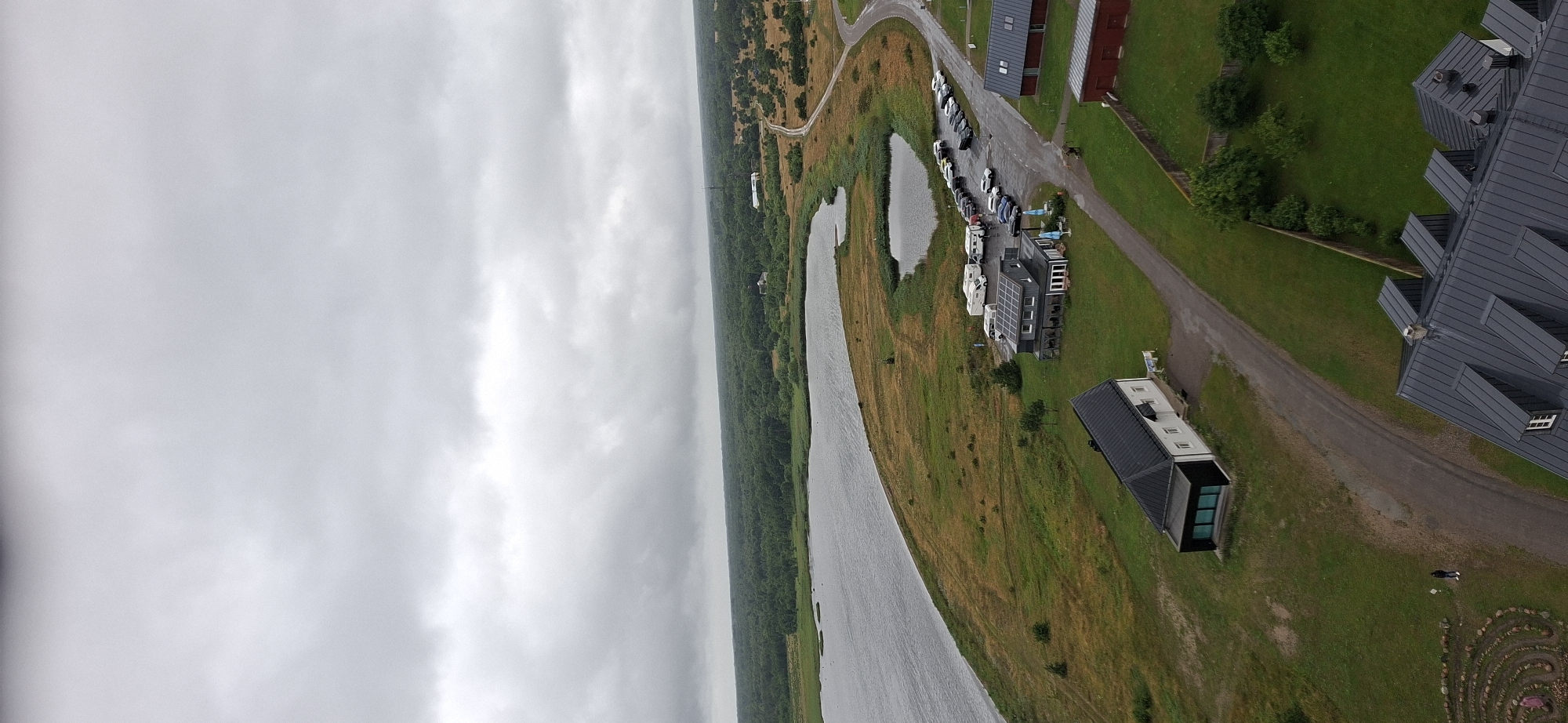 An aerial, vertical, medium shot captures an overcast day in Sääre, Estonia. The scene depicts a coastal landscape with a large body of water on the left, bordered by grassy terrain and a dense forest in the distance. Several buildings are visible, including a long, white structure with a black roof and a modern, dark building with solar panels. A parking lot with numerous vehicles, including cars and camper vans, is situated near the modern building. A road winds through the scene, connecting the buildings and parking area. A person is standing on a path near a circular stone maze. The overall atmosphere is serene and natural, with a hint of human activity in the form of buildings and vehicles.