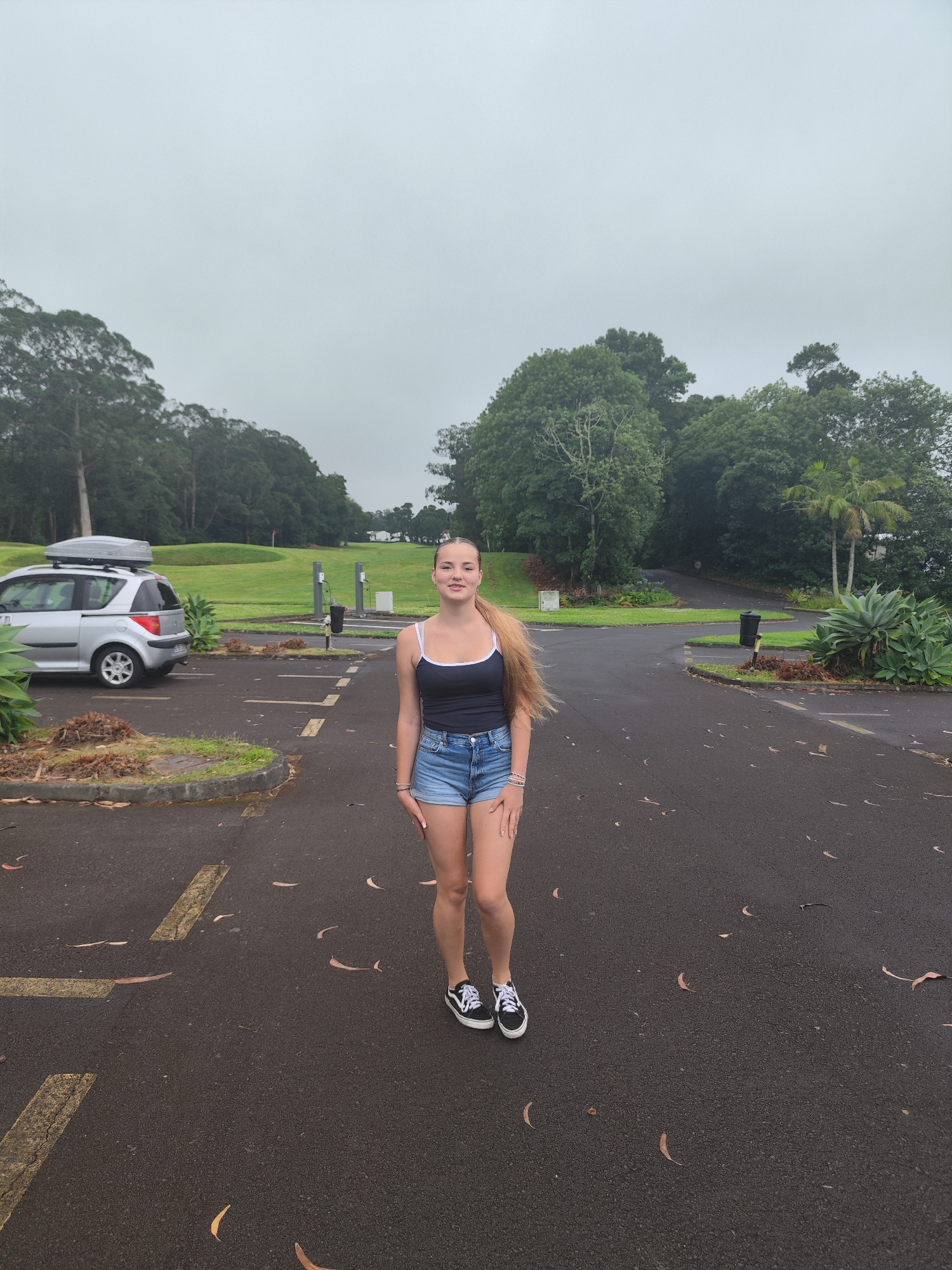 A young Caucasian female with long blonde hair and fair skin stands in the center of a paved area. She is wearing a black tank top with white trim, denim shorts, and black and white Vans sneakers. She is smiling at the camera. To her left, a silver station wagon with a roof rack is parked. The vehicle is facing left. Behind the woman, the paved area is marked with yellow lines, and there are planters with foliage. In the background, a golf course is visible with green grass and trees. The sky is overcast and gray. The location is an outdoor parking lot adjacent to a golf course, likely in Ponta Delgada, Portugal.