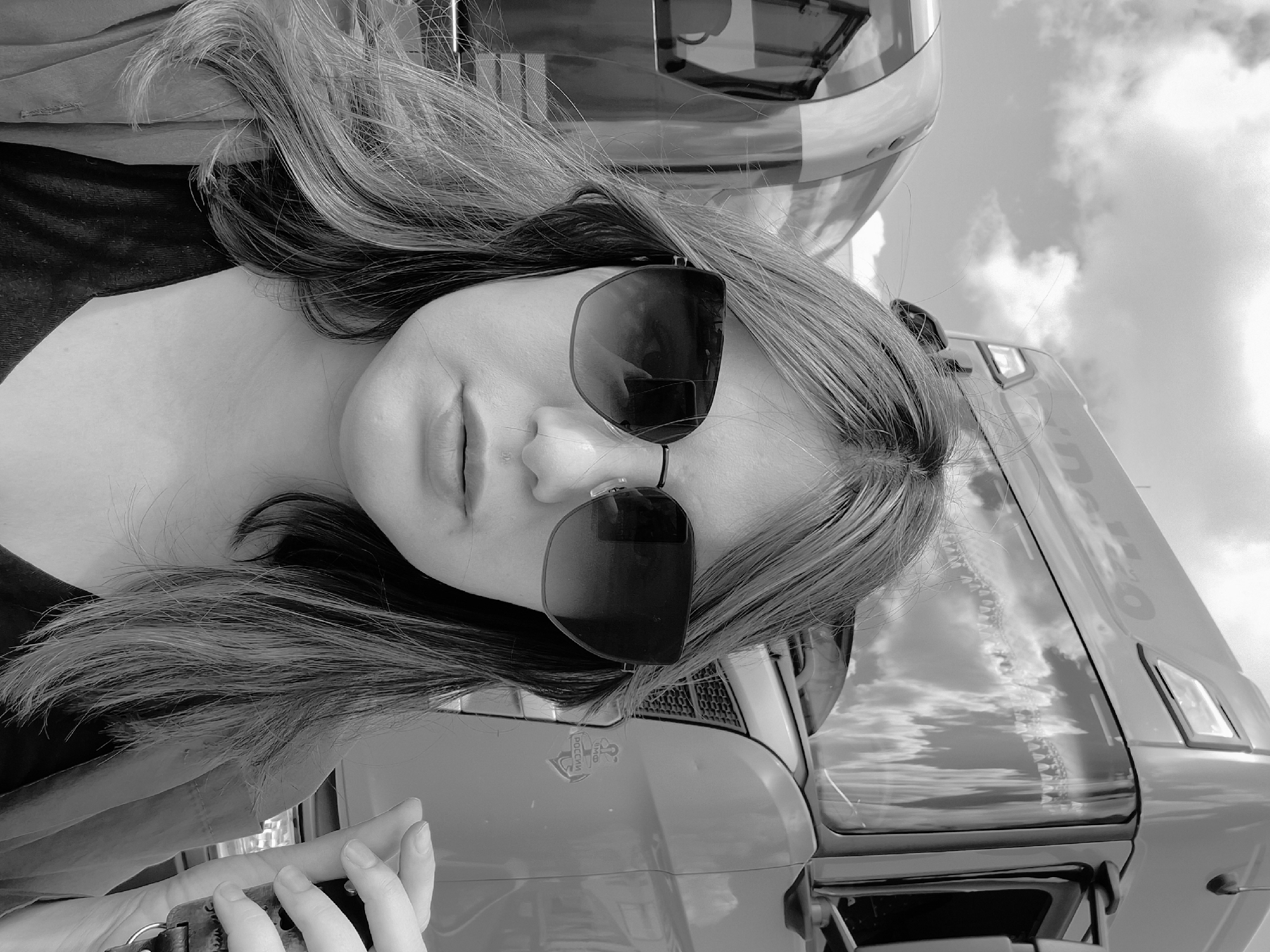 A black and white, vertically oriented outdoor portrait features a young woman positioned in front of a large truck, likely taken in Izhevsk, Russia. The woman, appearing to be in her late teens or early twenties, is the central subject, with her upper body visible. She has long, flowing hair with lighter streaks, parted on the side and draped over her shoulders. Large, dark-lensed sunglasses with thick frames obscure her eyes, and her lips are slightly parted in a neutral expression. She wears a dark top and a lighter jacket or shirt. Her left hand is visible in the lower left corner, holding a metallic clasp or strap, possibly from a bag.
The background is dominated by the large truck, which appears light-colored (possibly red or white in color). Its glossy surfaces and windows reflect the cloudy sky, suggesting a bright but overcast day. The truck's cabin structure, hood, and grille are visible around the woman. A notable detail is a round emblem on the truck's hood, clearly displaying the word "РОССИИ" (Rossii, meaning "of Russia" or "Russia's") and featuring a two-headed eagle, a national symbol. Below "РОССИИ", part of "ФЕДЕРАЦИИ" (Federatsii, meaning "Federation") is also discernible, explicitly confirming the location context of Russia. The overall mood is casual and observational. The image is originally oriented diagonally, 90 degrees counter-clockwise from a standard portrait view.