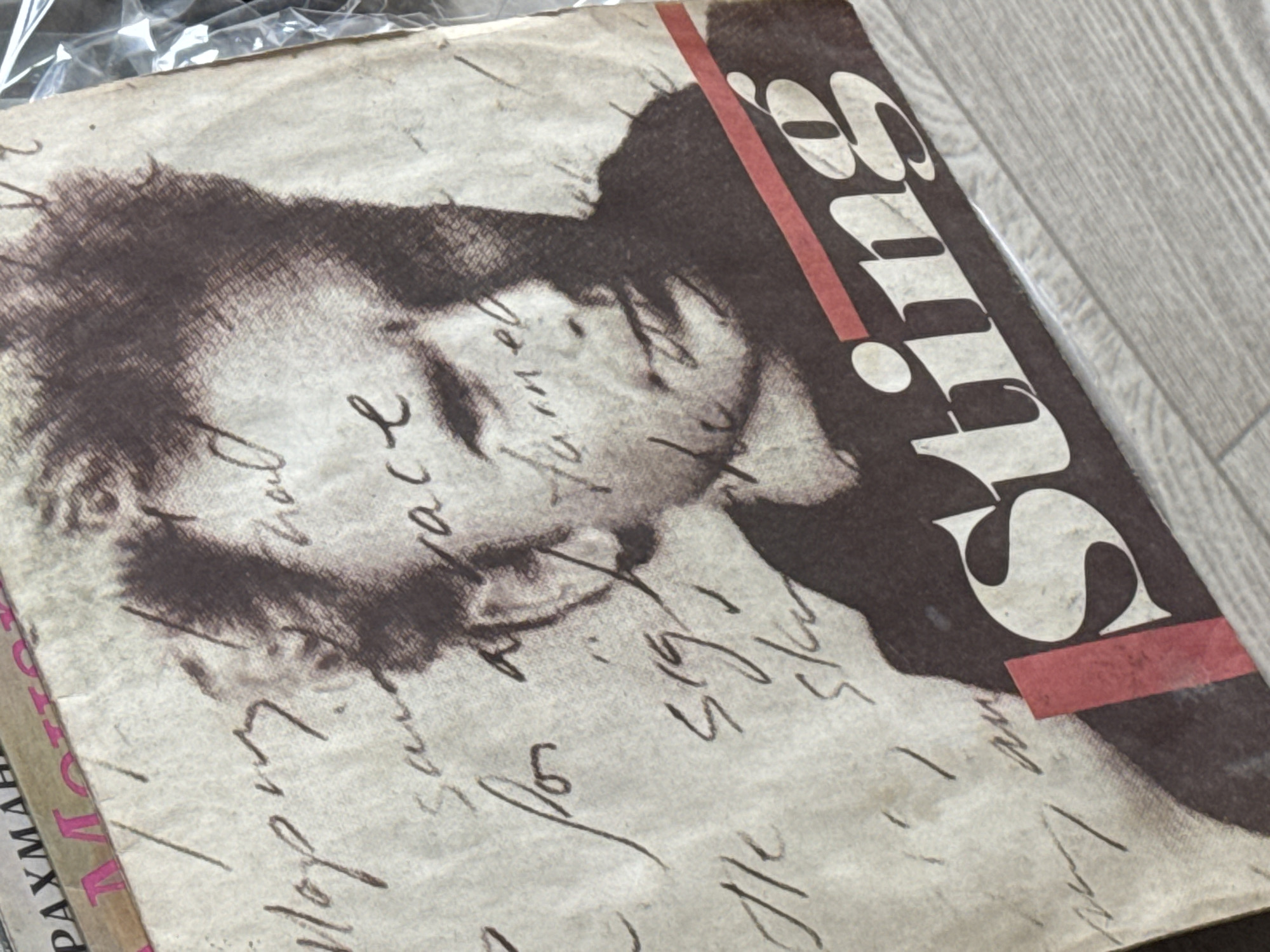 The image shows a weathered magazine cover. A faded, black and white image of a man's face occupies the left side of the cover, overlayed with handwritten text in what appears to be cursive. The word "Brits" is printed in bold, white font outlined in black down the right side of the cover. A red, rectangular stripe is overlaid diagonally across the letters. A partial word, "PAXMAMH" is visible along the left edge of the cover. The item is possibly sealed in a transparent plastic bag. The background appears to be a light-colored wooden surface. There is no information in the image to confirm it is taken in Kladovitsy, Russia.