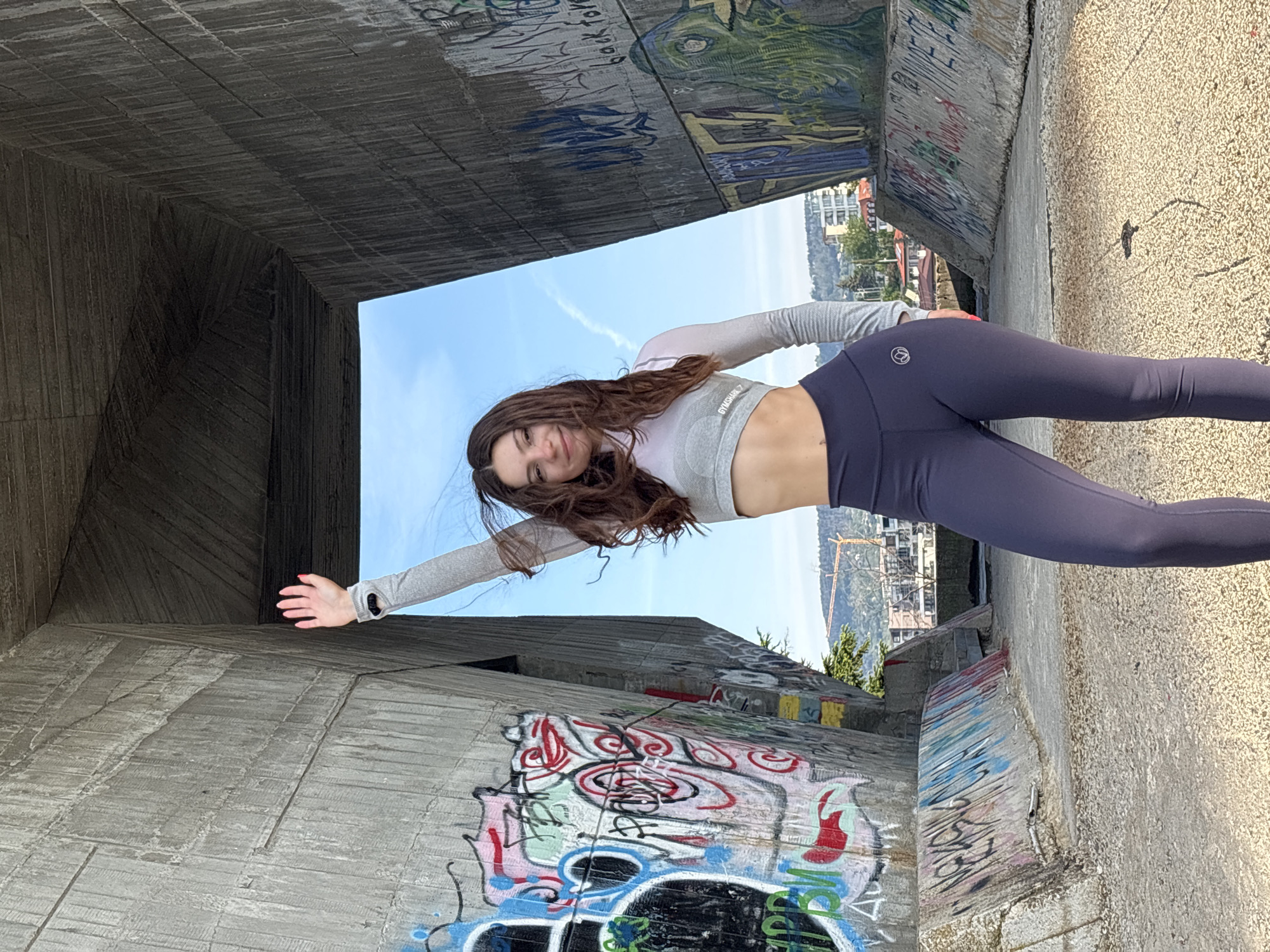 A young woman poses for a photo in what appears to be an urban underpass. The walls are made of concrete and are heavily covered in graffiti. The woman is wearing a light gray long-sleeved crop top and dark purple leggings. She is standing in a horizontal position, her right arm extended and her left arm near her body. Outside the underpass, there is a glimpse of buildings, trees, and a blue sky. It's unclear whether this is Varna, Bulgaria without additional information.