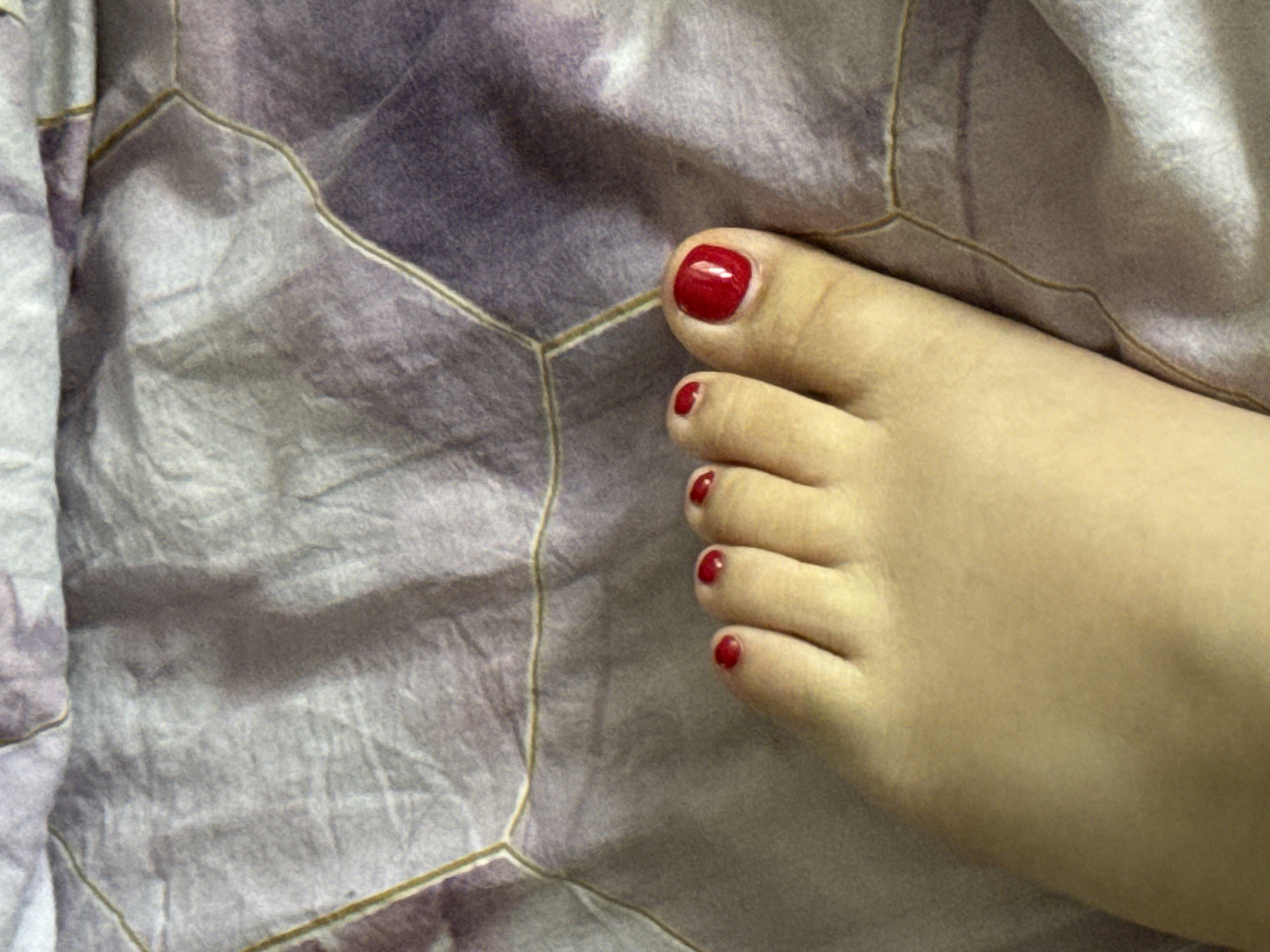 A close-up, overhead view shows a left foot with red nail polish resting on a textured fabric surface. The fabric has a pattern of light purple and white, with thin, golden lines creating a hexagonal or polygonal design. The foot is positioned in the lower right portion of the frame, with the toes pointing towards the top right. The skin tone of the foot is fair. The nail polish is a bright, glossy red. The lighting appears to be natural and diffuse, casting soft shadows. No specific location in Saratov, Russia can be determined from the image.