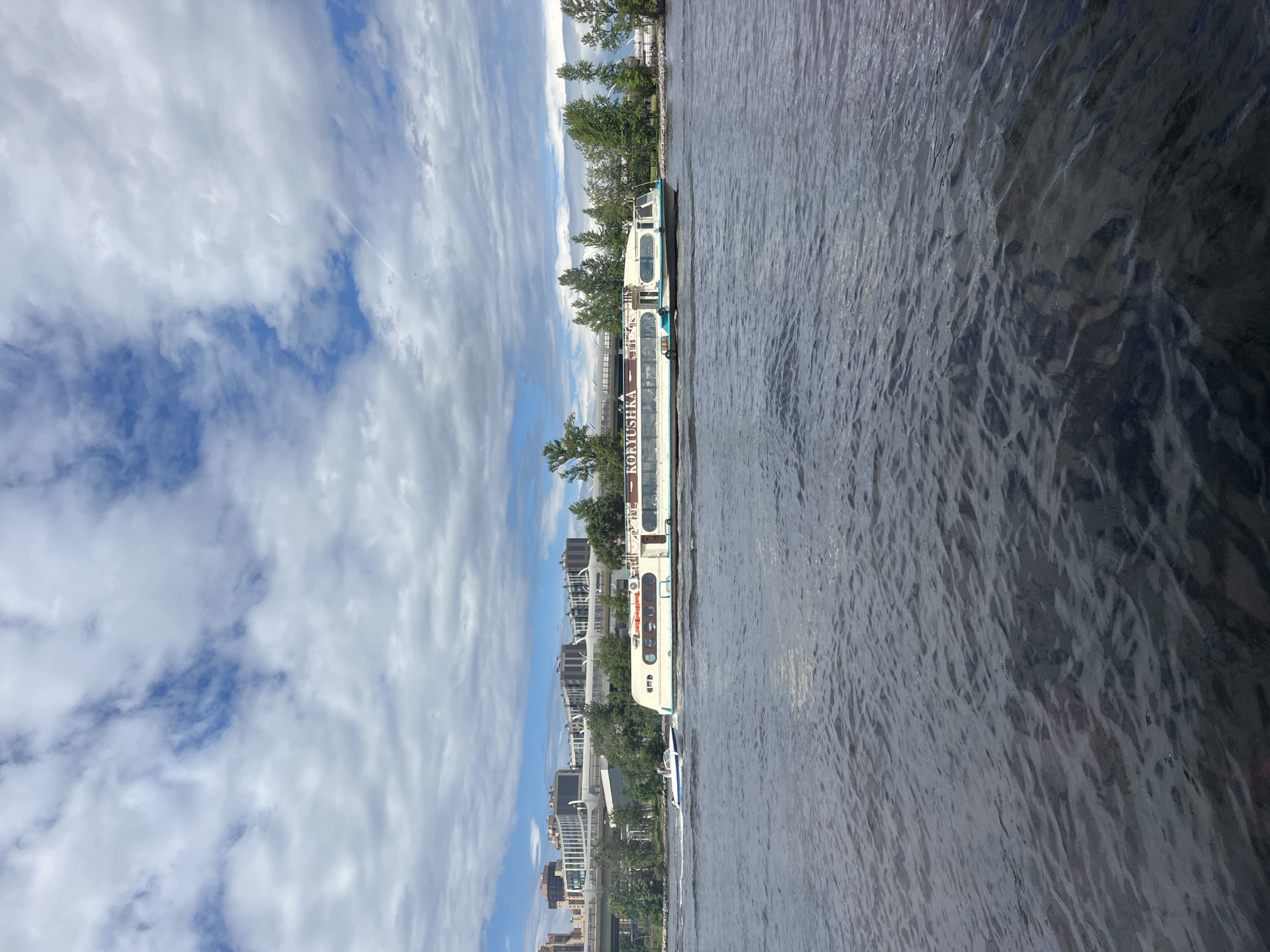 This media file is a photograph taken from a boat on a river in Saint Petersburg, Russia. The image is oriented vertically, with the sky occupying the left side and the water on the right.
The subjects include a white and teal passenger boat with multiple decks, labeled "KORYUSHKA" in red lettering. There are some people visible on the boat, and a smaller blue and white boat is trailing behind. The background shows a city skyline with modern buildings and trees lining the riverbank.
The scene is a daytime shot, likely in the late morning or early afternoon, given the bright but somewhat overcast conditions. The weather appears to be partly cloudy, with dramatic white clouds interspersed with patches of blue sky. The water is rippling, suggesting a gentle breeze. The location cues, such as the distinctive architecture and the river setting, strongly suggest Saint Petersburg. The overall mood is one of urban exploration and travel, with a sense of calm as the boat glides through the water.