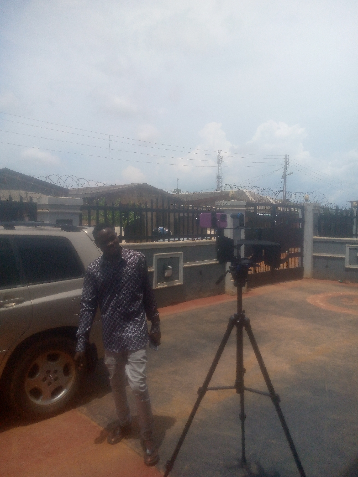 The image shows a man standing beside a silver SUV and a tripod holding cameras in Ikorodu, Nigeria. The man is wearing a patterned long-sleeved shirt and light-colored pants. The SUV is parked on a paved surface. Behind the tripod is a gated fence with barbed wire on top, surrounding a building. Power lines and a communications tower are visible in the background. The sky is partly cloudy.