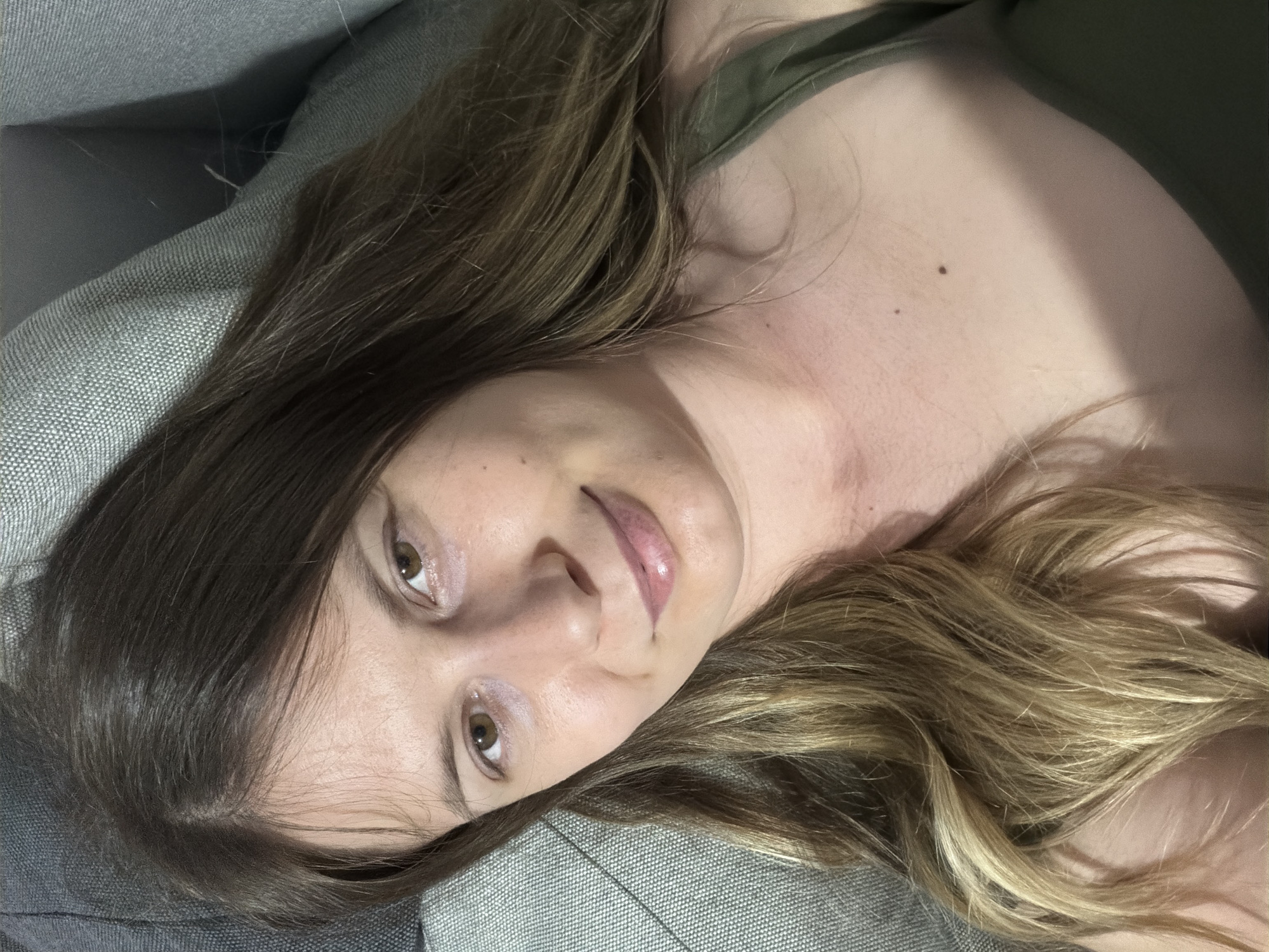 A close-up, overhead shot shows a woman with light brown, wavy hair that falls around her face and shoulders. She is lying down, appearing to be on a gray sofa, as indicated by the textured fabric visible in the background. The woman's head is tilted slightly, and she is looking directly at the camera with a gentle smile. Her eyes are a warm brown, and her lips are slightly parted, showing a hint of a smile. She is wearing a dark green tank top. The lighting suggests an indoor setting, possibly a home environment. There are no specific identifying landmarks or environmental features that would pinpoint a location in Stara Zagora, Bulgaria.