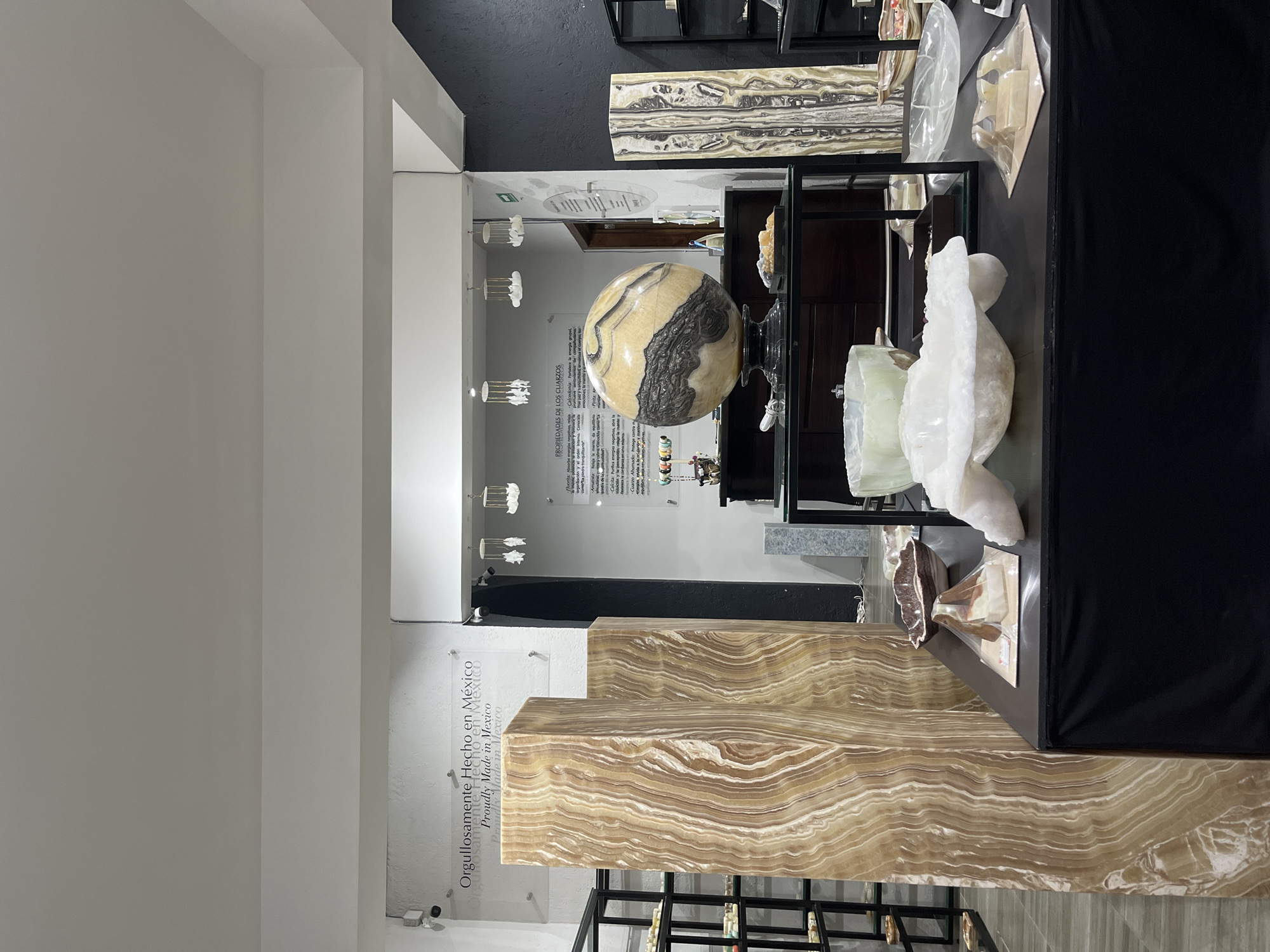 The image depicts an interior space, likely a showroom or retail establishment, in Domingo Arenas, Mexico, based on the "Orgullosamente Hecho en México" inscription. The scene features a display of what appears to be polished onyx or similar banded stone objects. These objects include a large sphere, rectangular blocks, and smaller carved pieces. In the background, there's informational text on a wall, possibly describing the properties of quartz. The lighting is a mix of natural and artificial, and the environment appears clean and modern. There are no people visible in the frame. The primary focus is on showcasing the stone artifacts.