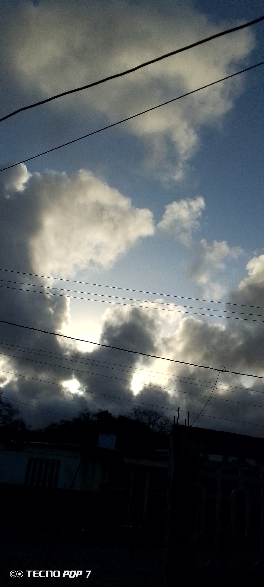 The image depicts a view of the sky in Altagracia, Venezuela. The sky is filled with cumulus clouds, some brightly lit by the sun, others dark gray and ominous. Power lines are visible across the sky, obscuring the view. Beneath the sky, there is a silhouette of a building and some trees. The image was taken with a Tecno Pop 7 phone.