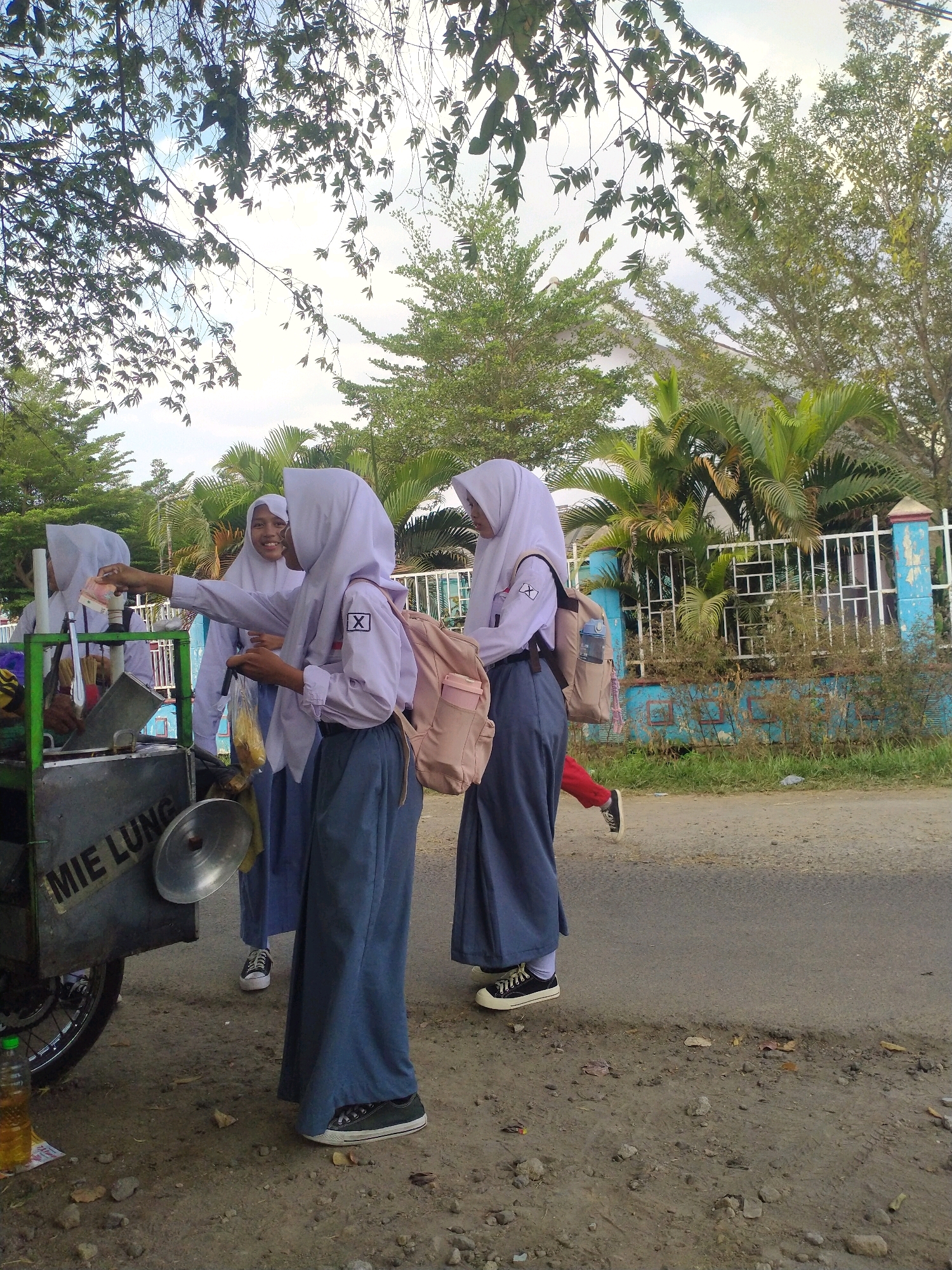 In Begajah, Indonesia, four young females, likely students based on their uniforms, are gathered around a mobile food cart. The cart, which reads "MIE LUNG," appears to be selling noodles. One student, wearing a white hijab, light purple shirt with an "X" emblem, and dark blue pants, is interacting with the cart, possibly making a purchase. Another student in a similar uniform is smiling and looking towards the first. A third student, also in uniform, is partially obscured. A fourth student, wearing a red top and dark pants, is walking away from the group. The scene takes place outdoors on a paved road, with trees, palm trees, and a light blue fence in the background. The sky is overcast.