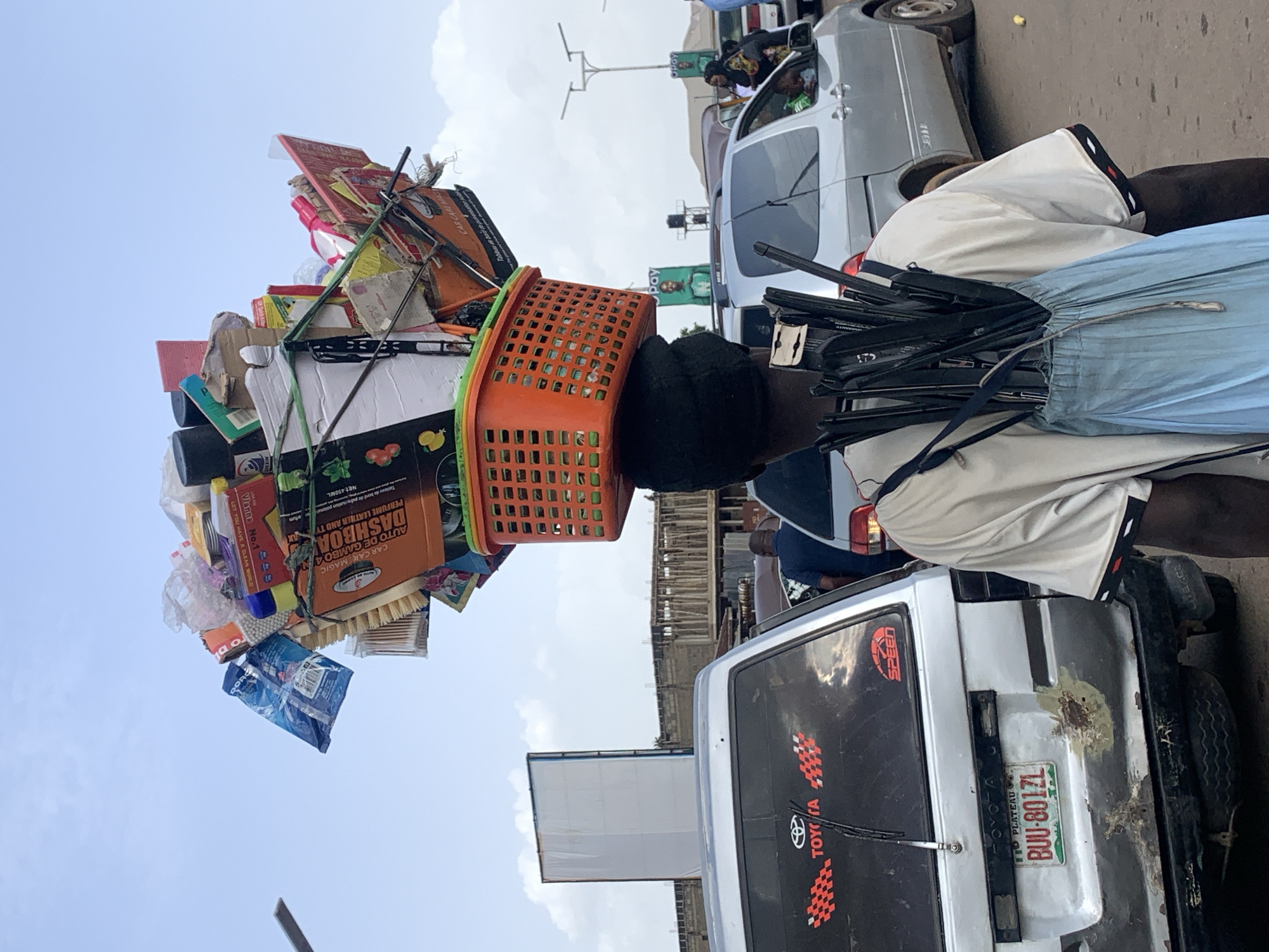 The image shows a person carrying a very large and precarious stack of goods on their head. The stack is composed of various items including boxes, bottles, a basket, and what appear to be car care products. The person is wearing a light-colored shirt and blue shorts. They are walking or standing on a street, with several vehicles visible in the background. One of the vehicles is a silver van with a "TOYOTA LITEACE" label and a license plate reading "BUU-801ZL" from Plateau State. The scene appears to be outdoors during the daytime, with a cloudy sky. The person's face is not clearly visible due to the angle and the items on their head. The overall impression is of a street vendor or transporter in Nigeria, likely in a busy urban environment. The text visible includes brand names and product descriptions such as "CAR CAR MAGIC," "DASHBOA," and "PERFUME LEATHER AND TY." The image is oriented vertically, suggesting it was taken with the phone held upright.