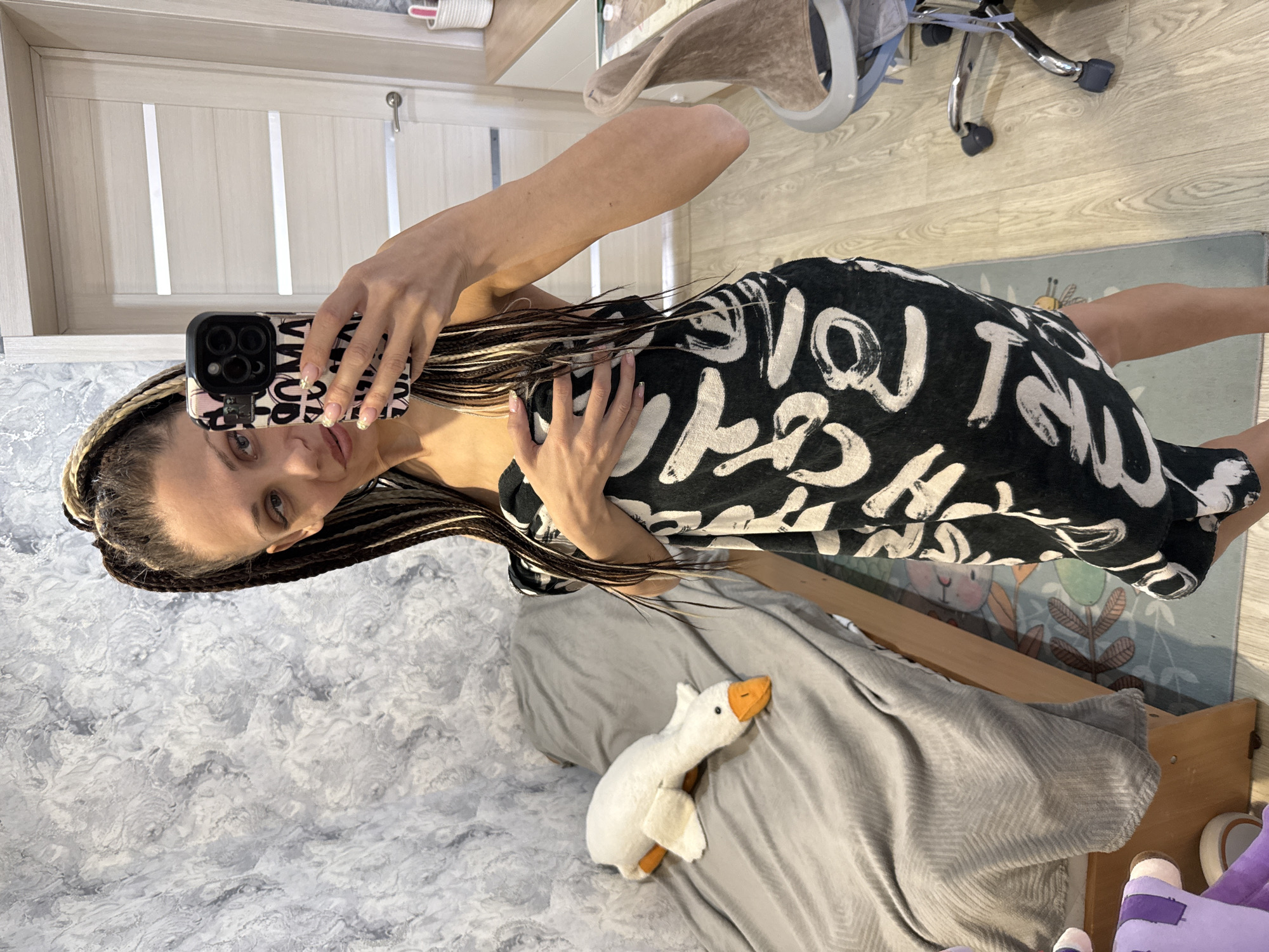 The image captures a woman taking a mirror selfie indoors in Rusanovo, Russia. She is positioned towards the center-right, looking directly into the camera with a neutral to slightly smiling expression. Her long hair is styled in two-toned braids (dark and blonde/light brown). She holds a black smartphone with a triple-camera module and a patterned case in her right hand; her fingernails are manicured with light polish. The phone case displays reversed text, appearing to be repetitions of "LOVE" or similar words.
She is wearing a black garment, likely a dress or large t-shirt, patterned with large, brush-stroke-style white letters that spell out words like "LOVE" or "YOU LOVE ME." Her left hand rests gently near her chest.
The indoor setting features a textured gray and white wall to her left and a light-colored wooden floor. Behind her is a bed covered with a gray blanket, where a white stuffed goose with an orange beak and feet rests. A wooden bed frame is visible, along with a light wooden door. On the floor to the right, a rug depicting a cartoon rabbit and plants is visible, along with the chrome base and wheels of an office chair. The ambient light suggests it is daytime.