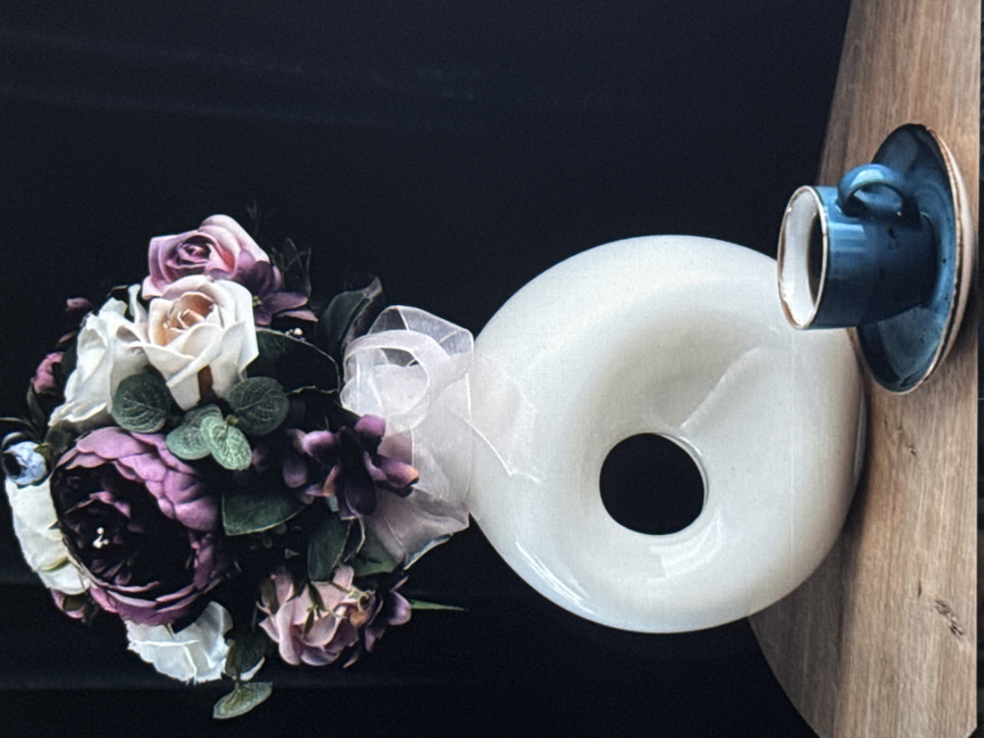 This image features a still life composition arranged on a wooden surface against a dark background. The primary subjects are a bouquet of artificial flowers and a small blue teacup and saucer. The bouquet is comprised of roses in various shades of pink, purple, and white, accented with green leaves and a sheer white ribbon tied around its base. To the right, a minimalist, white, rounded object with a central hole is positioned. A small, glossy blue teacup with a metallic rim rests on its matching saucer, which is also blue with a rustic metallic edge. The teacup is facing the viewer, appearing empty. The setting appears to be indoors, possibly a decorative arrangement for a cafe or home. The lighting is soft, creating gentle shadows. There is no visible text in the image. The overall mood is tranquil and aesthetically pleasing, highlighting the textures and colors of the objects. The composition is artfully arranged, suggesting a deliberate staging for a photograph.