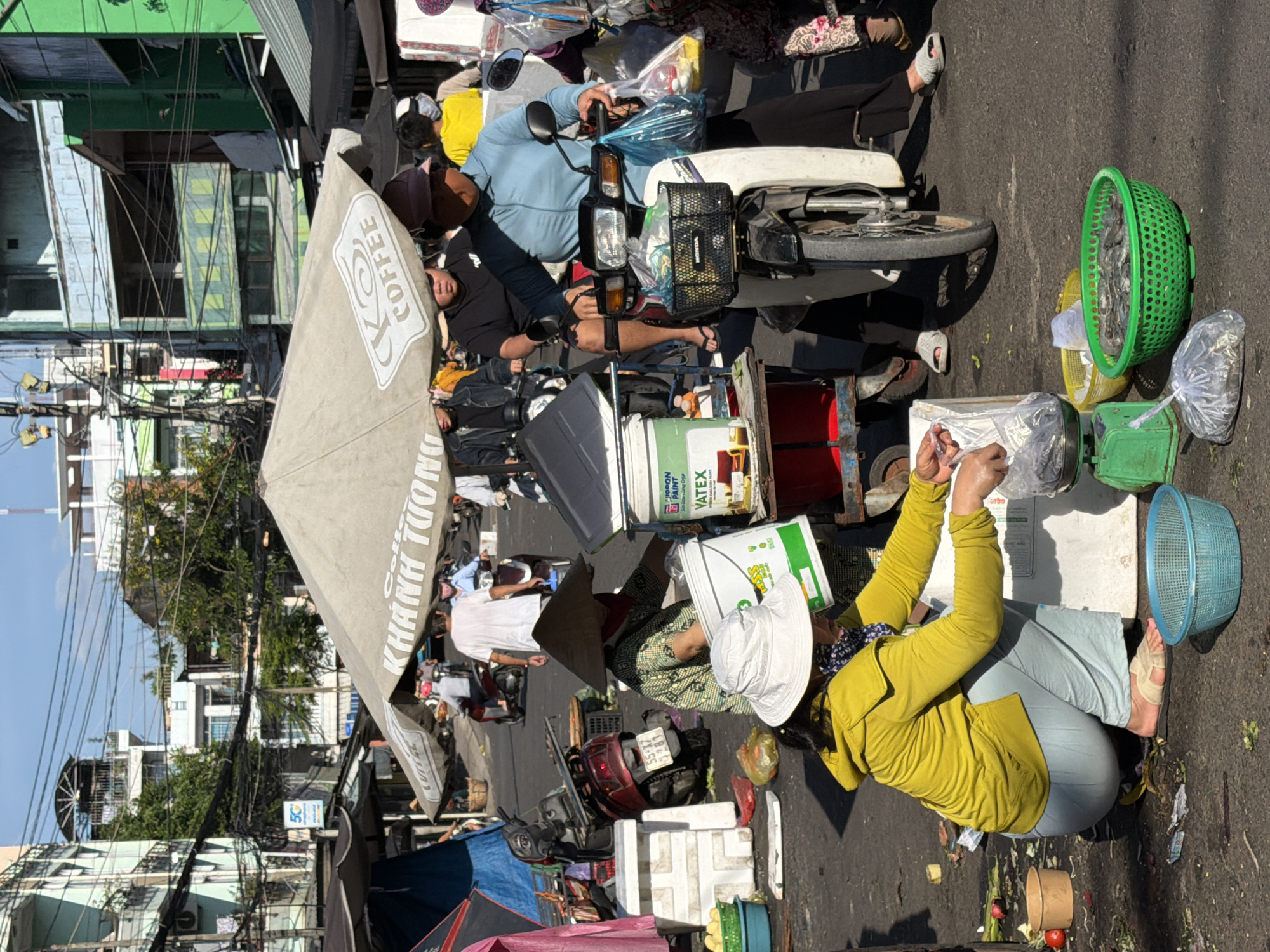 The image depicts a bustling street market scene in Nha Trang, Vietnam, under a bright, clear sky. The foreground is occupied by a woman in a yellow hoodie and light blue pants, crouching down and arranging fish in a green plastic basket. She wears a white bucket hat and a patterned blue and white scarf around her neck. To her left, a green digital scale is visible with a plastic bag of fish on it. Next to the basket of fish is a yellow bowl, and further to the left, a blue plastic basin.
Behind the woman, a large, light-colored umbrella provides shade, with the words "KHANH TUONG COFFEE" visible on it. Under the umbrella, several people are on motorcycles. One woman in a blue long-sleeved shirt and black pants is riding a white motorcycle, holding a blue plastic bag in her right hand and a white plastic bag in her left. Her face is partially obscured by a dark-colored hat. Another person in a black t-shirt is on a motorcycle behind her.
To the left of the umbrella, a red motorcycle is parked next to a white, modular cooler-like structure. A woman wearing a conical hat and a green patterned shirt is interacting with this structure. A red bucket and some food items are visible on the ground in front of her. Further left, a portion of a blue tarp-covered stall is visible, with pink fabric hanging down.
The background is filled with buildings, characteristic of a Vietnamese city, with balconies and visible electrical wiring crisscrossing overhead. Some buildings are painted in muted tones, while others are vibrantly colored green. Trees are interspersed among the buildings, adding to the urban foliage. The overall impression is one of a lively, everyday market day in Nha Trang.