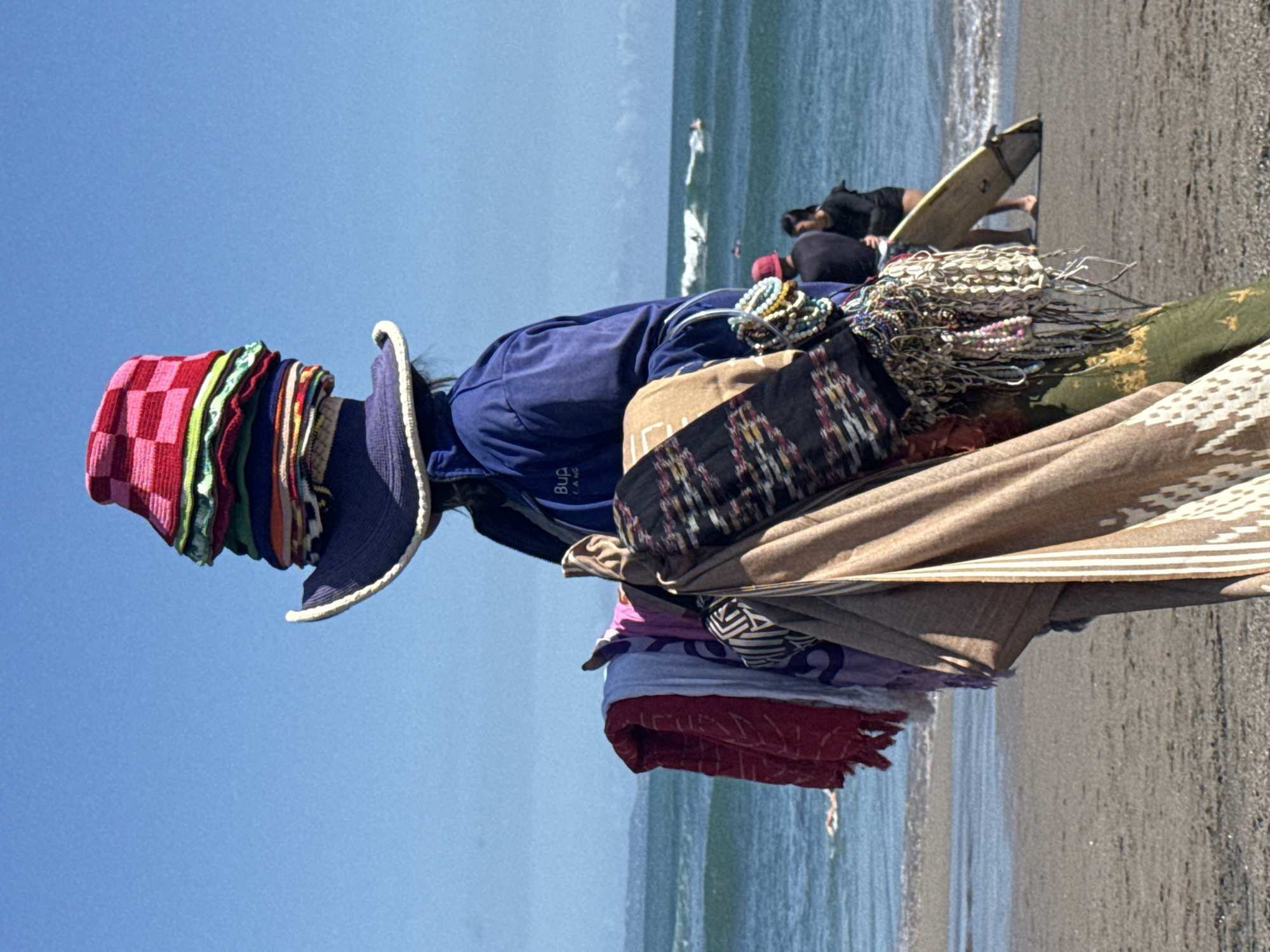The image shows a person on a beach, presumably a vendor. The person is heavily laden with merchandise, including a stack of hats on their head, a variety of bracelets and necklaces, and draped fabrics. The vendor is standing near the shoreline on the beach. In the background, people are visible, one lying on a surfboard. The location is North Kuta, Indonesia, suggested by the beach scene.