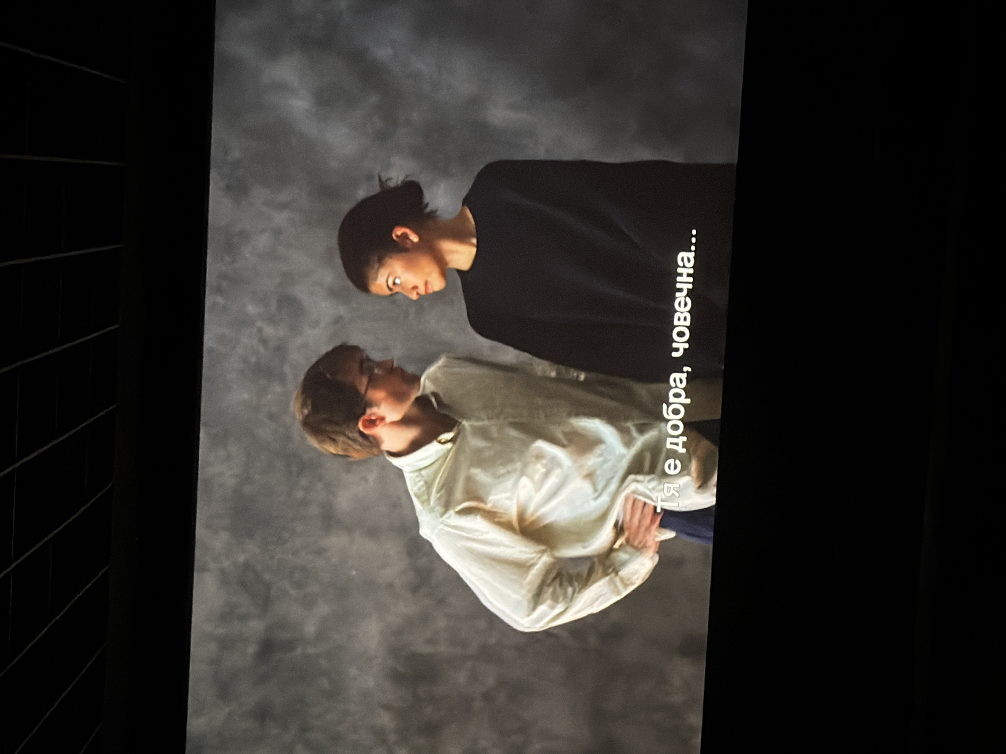 The image, captured in Sofia, Bulgaria, displays two individuals in a close-up, possibly a scene from a film, play, or a staged photoshoot. The background is a mottled grey with a soft, textured appearance, suggesting an indoor studio or stage setting. The overall lighting is even, creating a contemplative atmosphere.
On the left, a male with light brown hair is seen wearing a light-colored, possibly cream or off-white, long-sleeved button-up shirt over a darker layer. He looks upwards towards the other person, with a gentle, focused expression. His right hand is visible, casually tucked into his left arm or waist area.
Facing him on the right is a female with dark hair, pulled back, wearing a dark, long-sleeved top. Her gaze is directed downwards towards the male, and her expression suggests concentration or introspection, with a slight furrow to her brow.
The two subjects are positioned intimately, facing each other, implying a moment of deep conversation, connection, or shared emotion. The scene conveys a serious and perhaps tender interaction.
Partially visible white text is superimposed on the dark background to the right of the female. When viewed correctly, the legible portion reads: "Тя е добра, човечна...", which translates from Bulgarian to "She is good, humane...". This text adds a narrative layer, possibly alluding to the character or theme depicted.