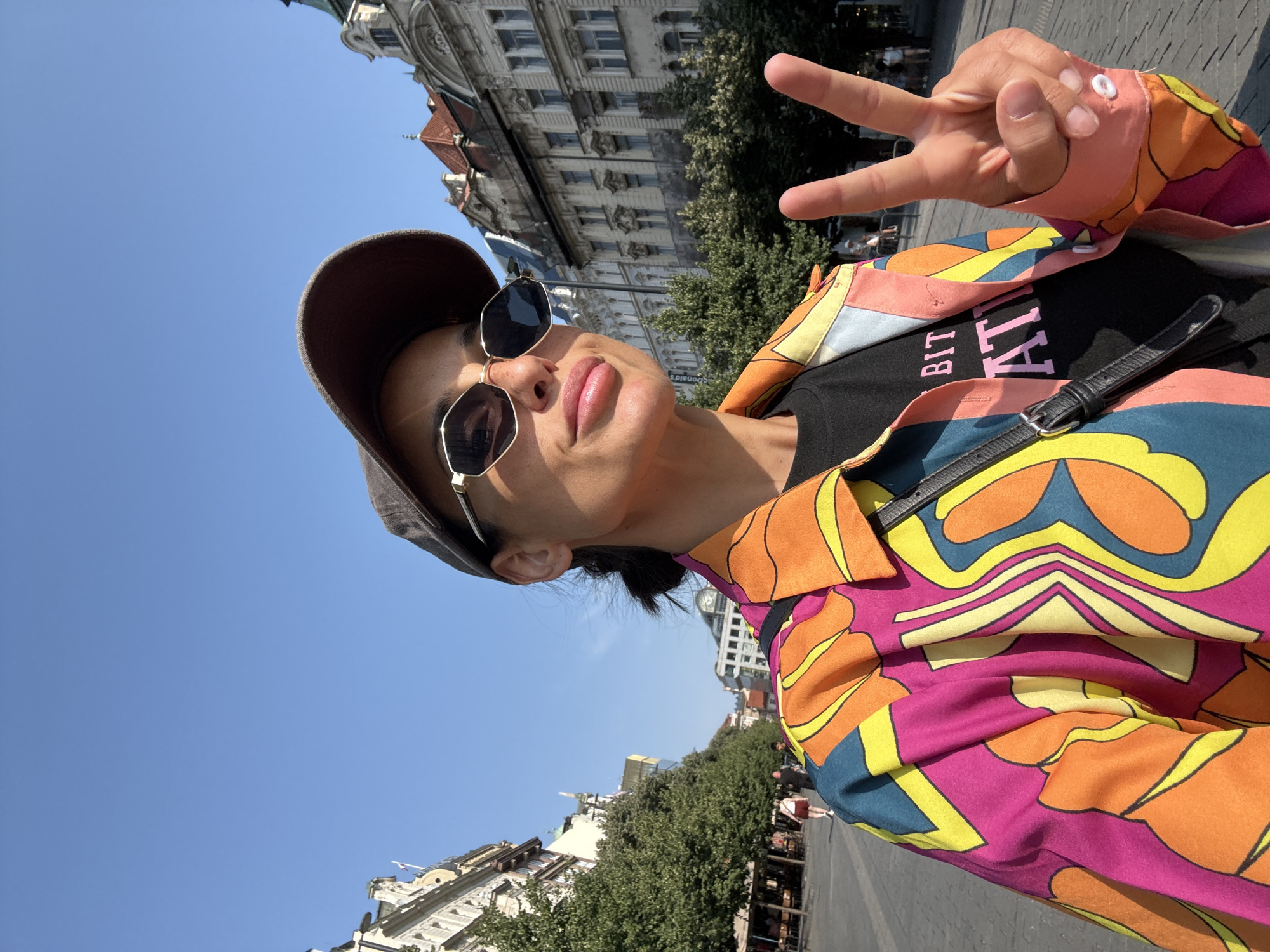 A woman wearing a colorful jacket, sunglasses, and a baseball cap takes a selfie in an outdoor public square. She is holding up two fingers in a peace sign. In the background, there are historic buildings, trees, and people walking. The scene appears to be in Prague, Czechia, given the architectural style of the buildings and the cobblestone pavement. The woman's t-shirt has text on it that is partially obscured.