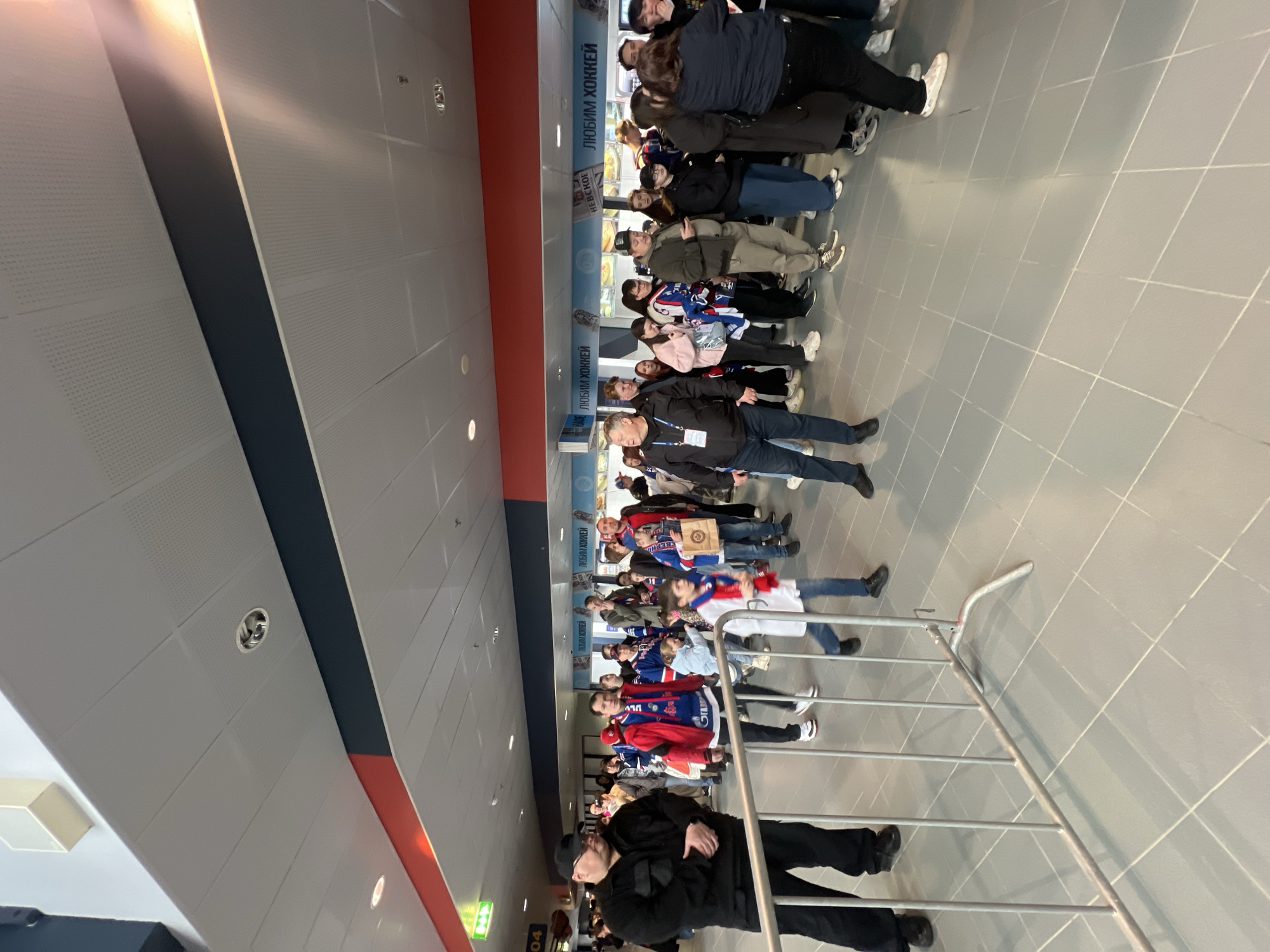 The image shows a queue of people in an indoor space. They are lined up along a grey tiled floor, separated from the viewer by a silver railing. Many of the people are wearing clothing associated with the "SKA" hockey team of St. Petersburg, Russia, including jerseys and scarves with the team's colors and logo. There is a sign along the wall behind the people that includes the phrase "любим хоккей," which translates to "we love hockey." The ceiling has a white, perforated panel design, with a wide stripe of black and a stripe of red along it. A sign showing the letter A with arrows around it is also visible in the foreground. Based on the hockey team clothing, the location is most likely within or near the Ice Palace in Saint Petersburg, Russia.
