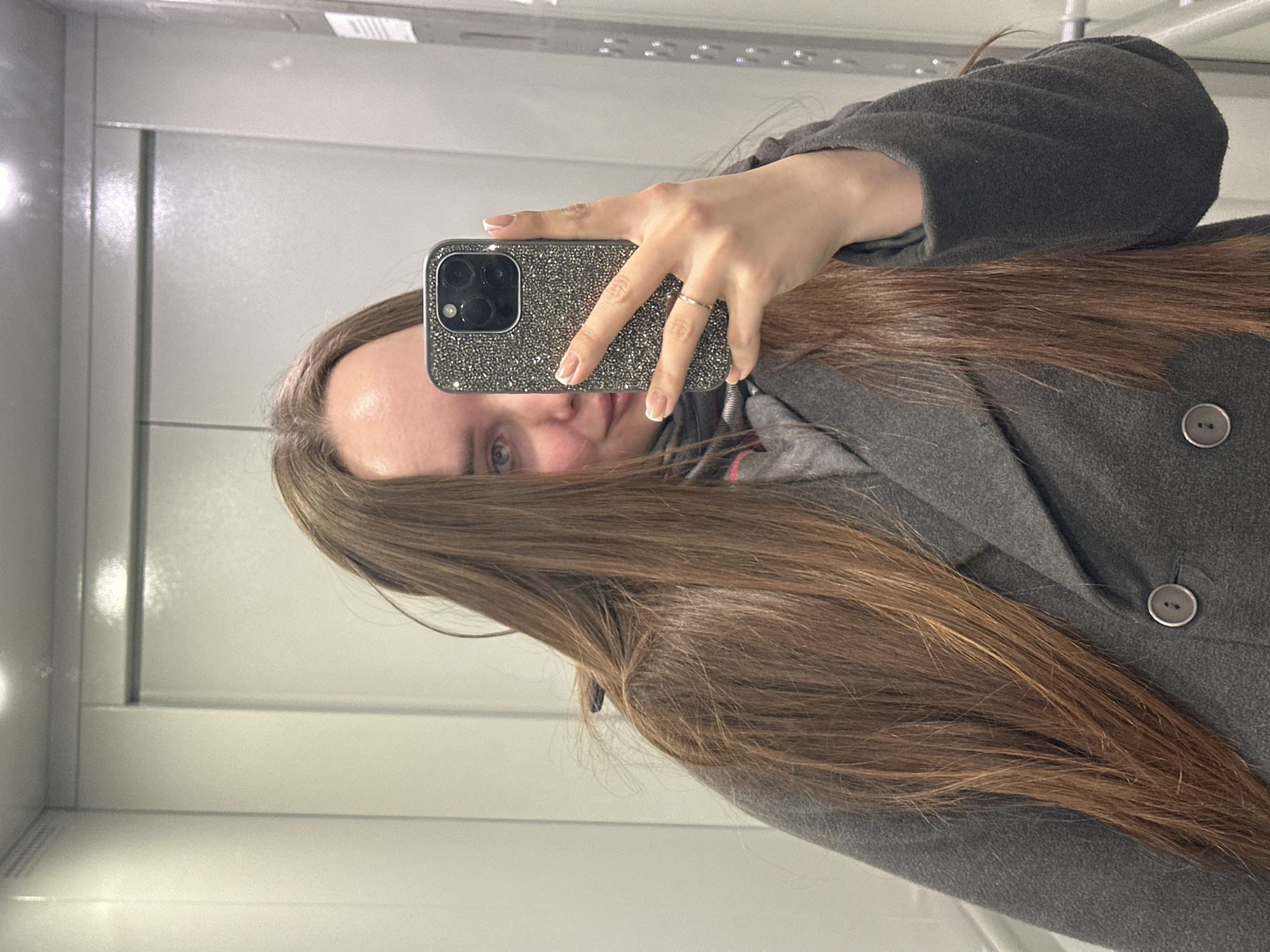 A woman with long brown hair takes a selfie in a mirrored enclosure, likely an elevator. She wears a grey coat with buttons and a grey hoodie underneath. Her phone, with a studded case, partially obscures her face. The background is a plain white wall with horizontal panels. The location appears to be indoors, possibly in an elevator. The city of Chaikovsky, Russia, cannot be determined from the image.
