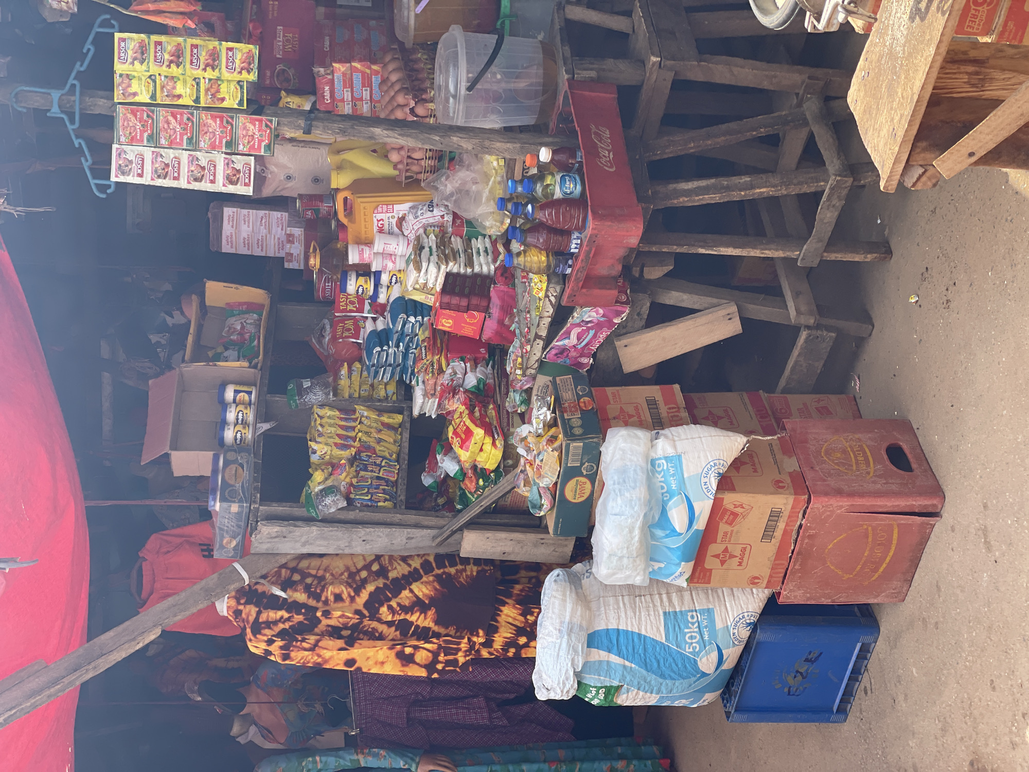 The image depicts an outdoor market stall in Akwanga, Nigeria, with a variety of goods for sale. The stall is covered by a red tarp, and wooden structures support the display shelves. Products include bags of flour or sugar, boxes of Maggi seasoning, bottles of drinks, cans of food, packets of spices, eggs, and cooking oil. There are also clothing items displayed, including a vibrant tie-dye patterned garment. The ground is paved with concrete. The scene appears to be a typical market environment with a focus on food and household items.