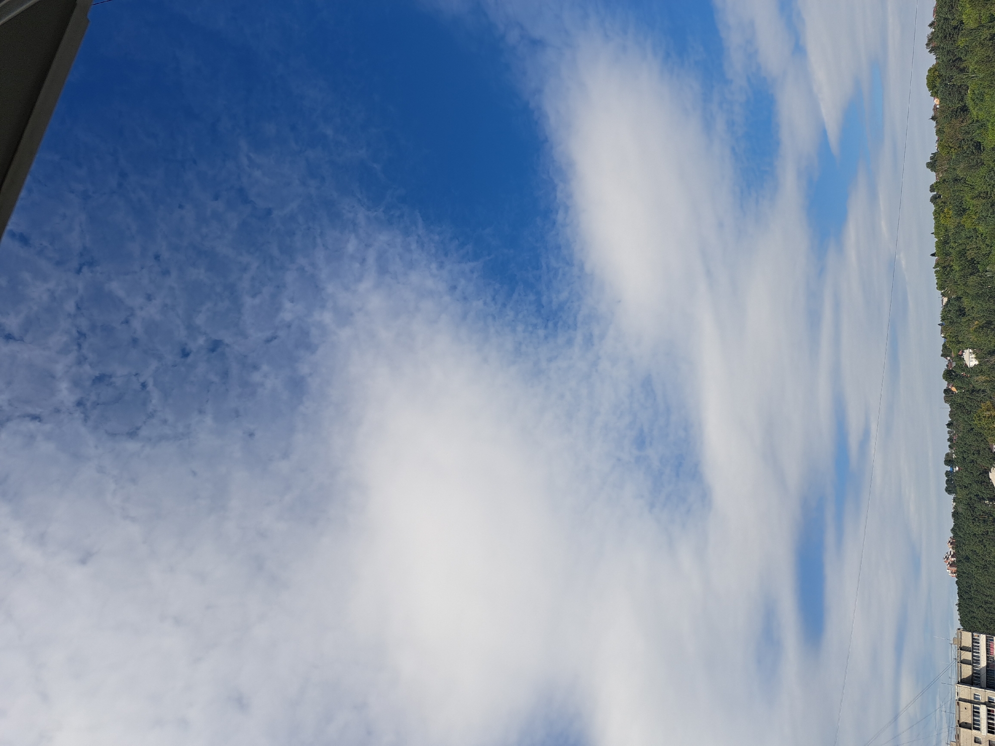 The image shows a bright blue sky with scattered white clouds, some thin and wispy, others more dense and fluffy. The top edge of the frame is partially obscured by a dark gray overhang, possibly part of a building or roof. Below the sky, a dense, green treeline stretches across the horizon, with a few buildings peeking through the foliage. The buildings are mostly residential, with visible windows and balconies. The overall scene suggests a clear day with moderate cloud cover, likely taken from an elevated position within the city of Irkutsk, Russia.