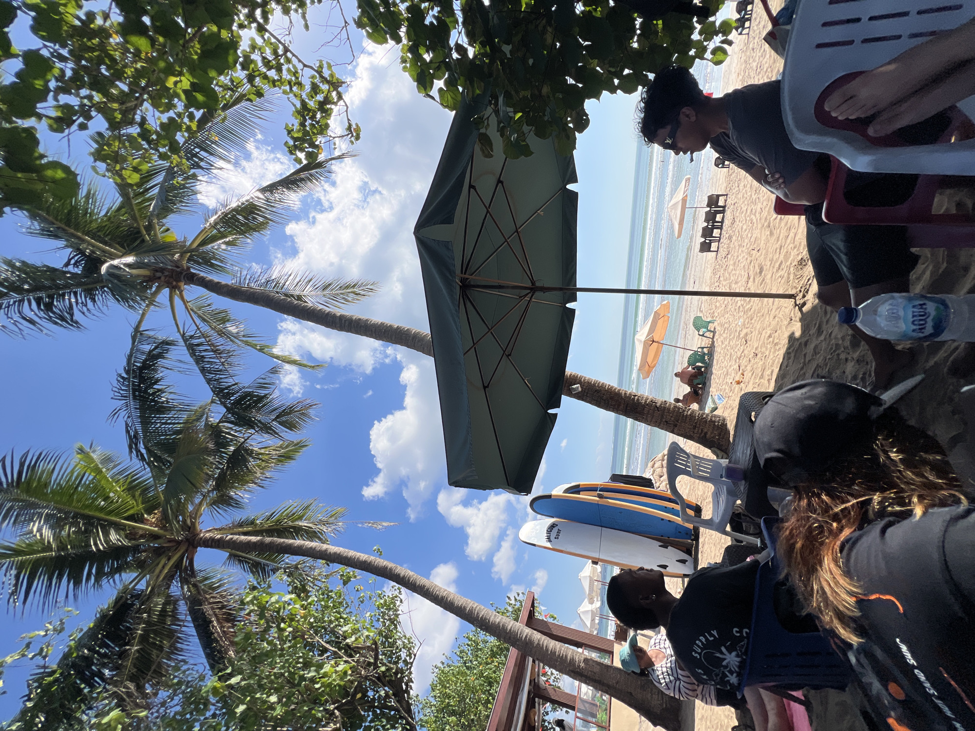 In Kuta, Indonesia, at a beachfront cafe, several people relax on a sandy shore under the shade of palm trees and a large green umbrella. In the foreground, a person wearing a black t-shirt and a black baseball cap faces away from the camera, with another person's head visible in the lower right corner. To the left, a person wearing a black t-shirt with a white graphic print is seated in a blue chair. Next to them, another person in a striped shirt and a blue sun hat is also seated.
Behind these individuals, several surfboards are propped up, leaning against a wooden structure, two of which are white and blue. White plastic chairs are scattered around the sand. In the midground, the calm ocean stretches towards the horizon, with small waves lapping the shore. Two orange umbrellas are open on the beach, with people sitting in chairs underneath them. Further back, more beach chairs and umbrellas are visible, suggesting a popular relaxation spot. The sky is a clear blue with scattered white clouds. The overall scene depicts a leisurely day at the beach.