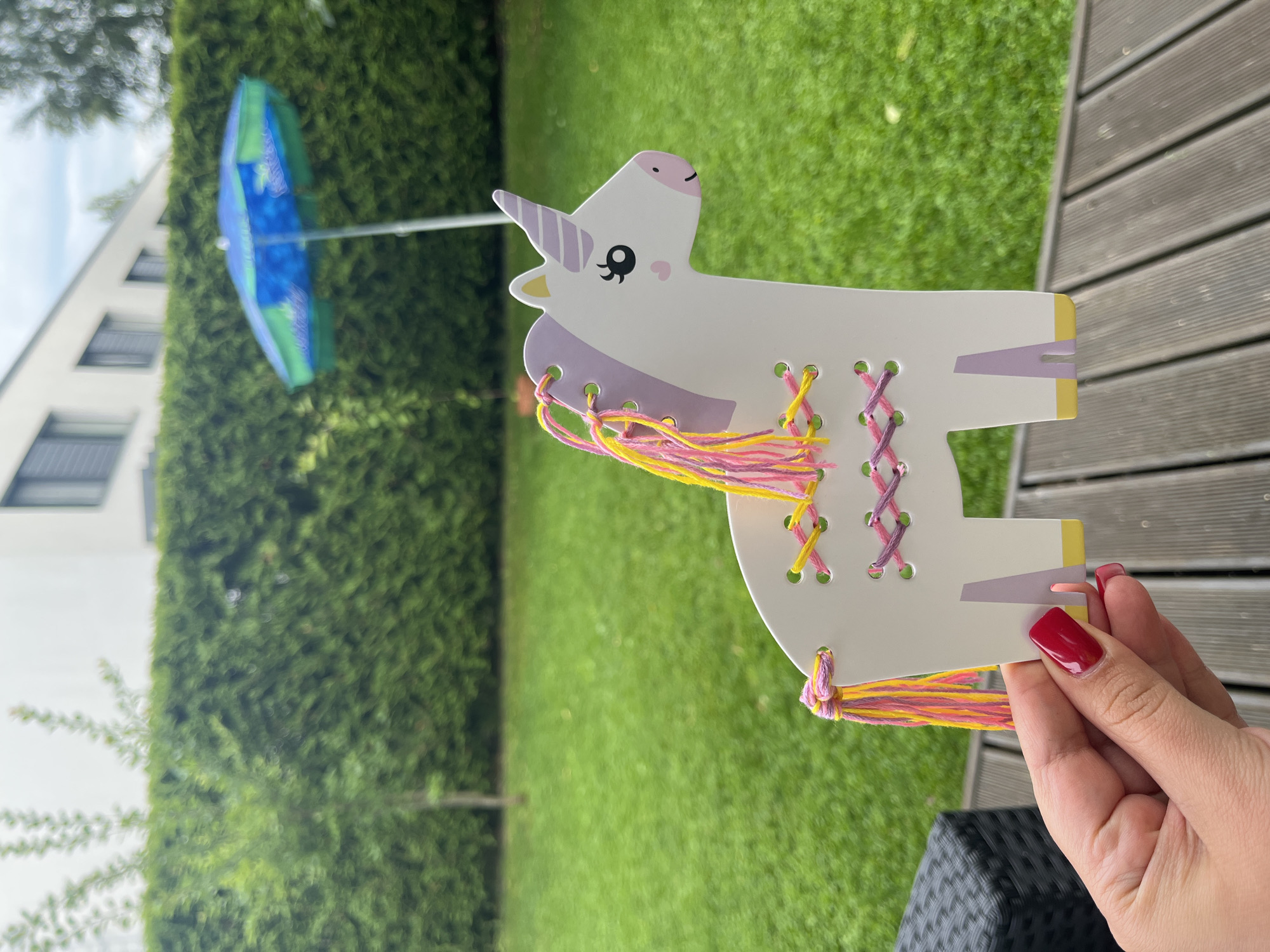 A person's hand with red-painted fingernails holds a white unicorn craft with colorful yarn threaded through holes in its body and tail. The unicorn has a purple horn, black eyes, and pink cheeks. The background shows a green lawn, a hedge, a building with windows, and a blue and green umbrella. The scene appears to be a backyard in Haabneeme alevik, Estonia, possibly on a cloudy day.