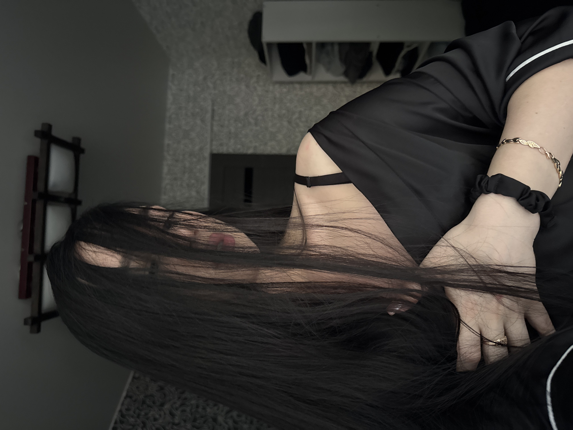 A young woman with long, dark hair is partially obscured by her hair, her face turned away from the camera. She is wearing a black silk robe with white trim and a black bra. A gold ring is on her left hand, and a black scrunchie and a gold bracelet are on her right wrist. The setting appears to be a bedroom with textured wallpaper and a shelf unit visible in the background. The image was taken in Novosibirsk, Russia.