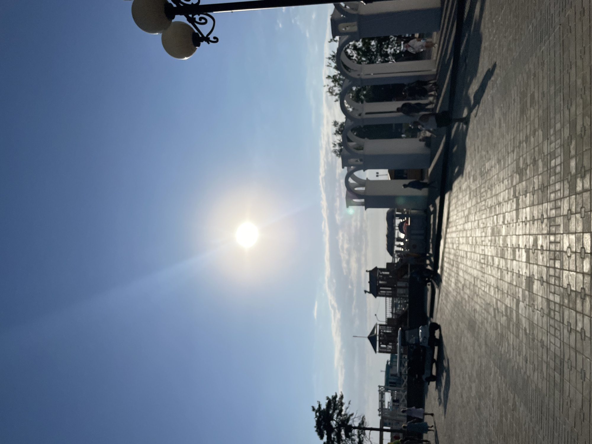 The image depicts a bright, sunny day in Anapa, Russia, with a clear blue sky and a prominent sun casting long shadows. The scene appears to be a waterfront promenade or a park.
In the foreground, a lamppost with two globes is visible at the top left. The ground is paved with light-colored tiles, creating a textured surface. The sun's rays are reflecting strongly off the pavement, indicating it is likely midday or early afternoon.
To the right of the frame, an arched structure, possibly part of a gazebo or a decorative walkway, extends along the promenade. Several people are scattered throughout the scene, some walking, others possibly sitting or standing near the arched structure. The attire of the people suggests warm weather.
Further into the background, there are glimpses of what appear to be small structures, perhaps kiosks or playgrounds, and trees. The overall impression is of a public space being enjoyed on a pleasant day. The orientation of the image is on its side, rotated 90 degrees clockwise from a typical upright view.