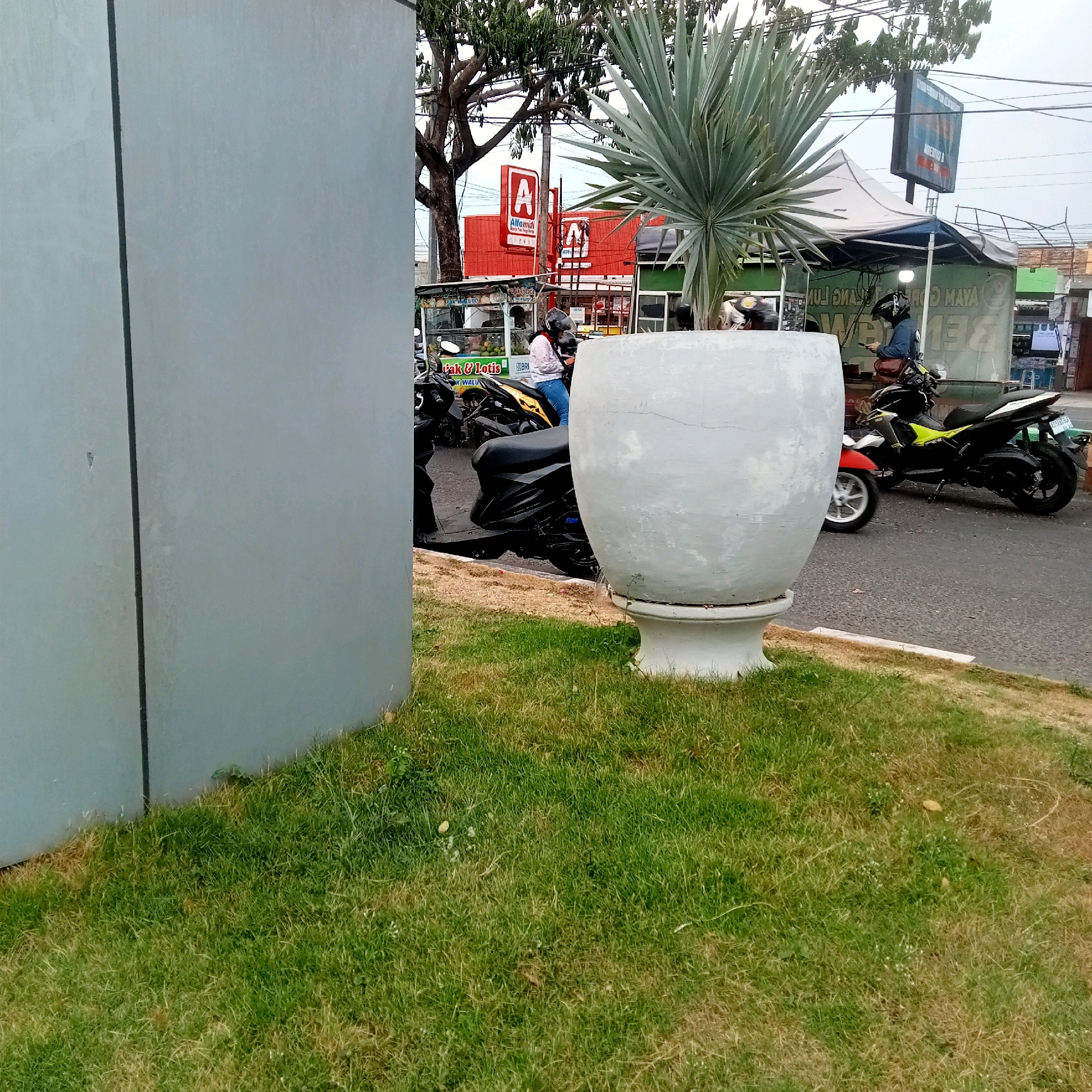 In Karanganyar, Indonesia, a street scene unfolds with a focus on a large, light gray decorative planter in the foreground, positioned on a patch of green grass. Behind the planter, a paved road is visible with several motorcycles parked and in motion. To the left, a tall, gray wall with a vertical dark line dominates the frame, partially obscuring the background. Further back, a bright red building with prominent "A" logos, identified as an Alfamidi convenience store, stands out. In front of the store, a small food stall is set up. Various people, mostly wearing helmets, are seen on or near the motorcycles. Trees with lush green foliage are interspersed throughout the scene, and power lines stretch across the overcast sky.