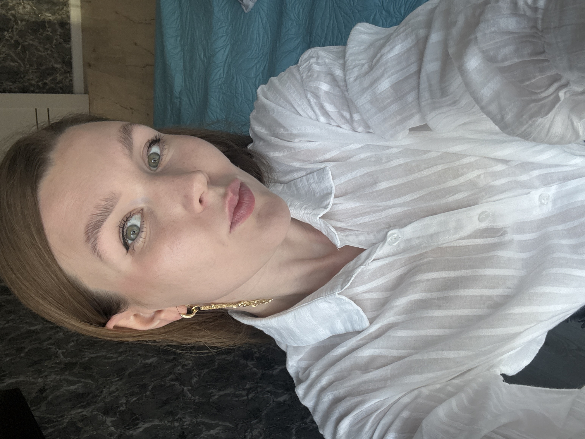 A young woman with fair skin, green eyes, and light brown hair is taking a selfie. She is wearing a white, vertically striped, sheer button-down shirt and a long gold earring. Her lips are pursed, and she is looking at the camera. The background is out of focus, but appears to be a dark, textured wall on the left and a textured blue surface on the right, possibly a bedspread. The image is taken indoors, in what appears to be a bedroom. There is no indication of the specific location within Izhevsk, Russia.
