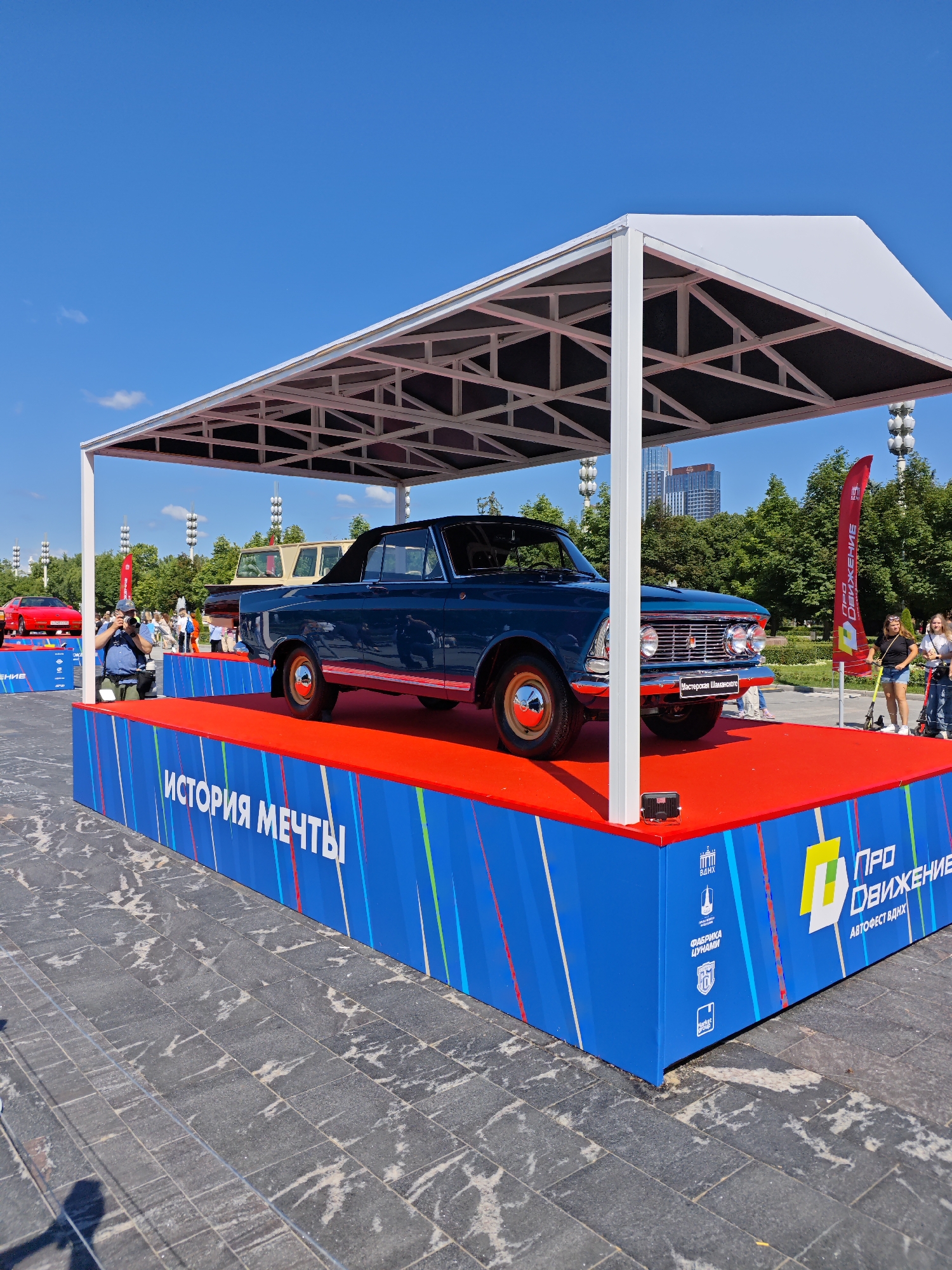 An outdoor automotive exhibition is taking place under a white canopy structure. A dark blue convertible is prominently displayed on a raised red platform, which is surrounded by a blue base featuring colorful stripes and text. The text on the blue base reads "ИСТОРИЯ МЕЧТЫ" (History of Dreams) and other organizational logos and text related to "ВДНХ" (Exhibition of Achievements of National Economy) and "АВТОФЕСТ ВДНХ" (Autofest VDNKh). In the background, a red sports car is visible on another platform, along with trees, streetlights, and buildings, suggesting an urban park setting. Several people are present, some observing the cars and others participating in the event, including a photographer with a camera. The sky is clear and blue, indicating fair weather. The exhibition appears to be located at VDNKh in Moscow, Russia, given the prominent VDNKh branding.